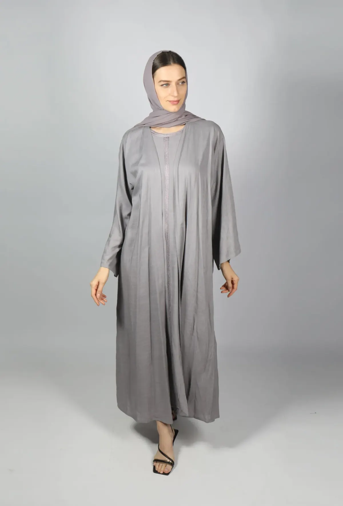 Perempuan memakai abaya abu-abu terbuka di atas gamis dalaman abu-abu, jilbab pashmina ungu keabuan, dan sandal hak tali hitam, berpose di studio berlatar abu-abu.