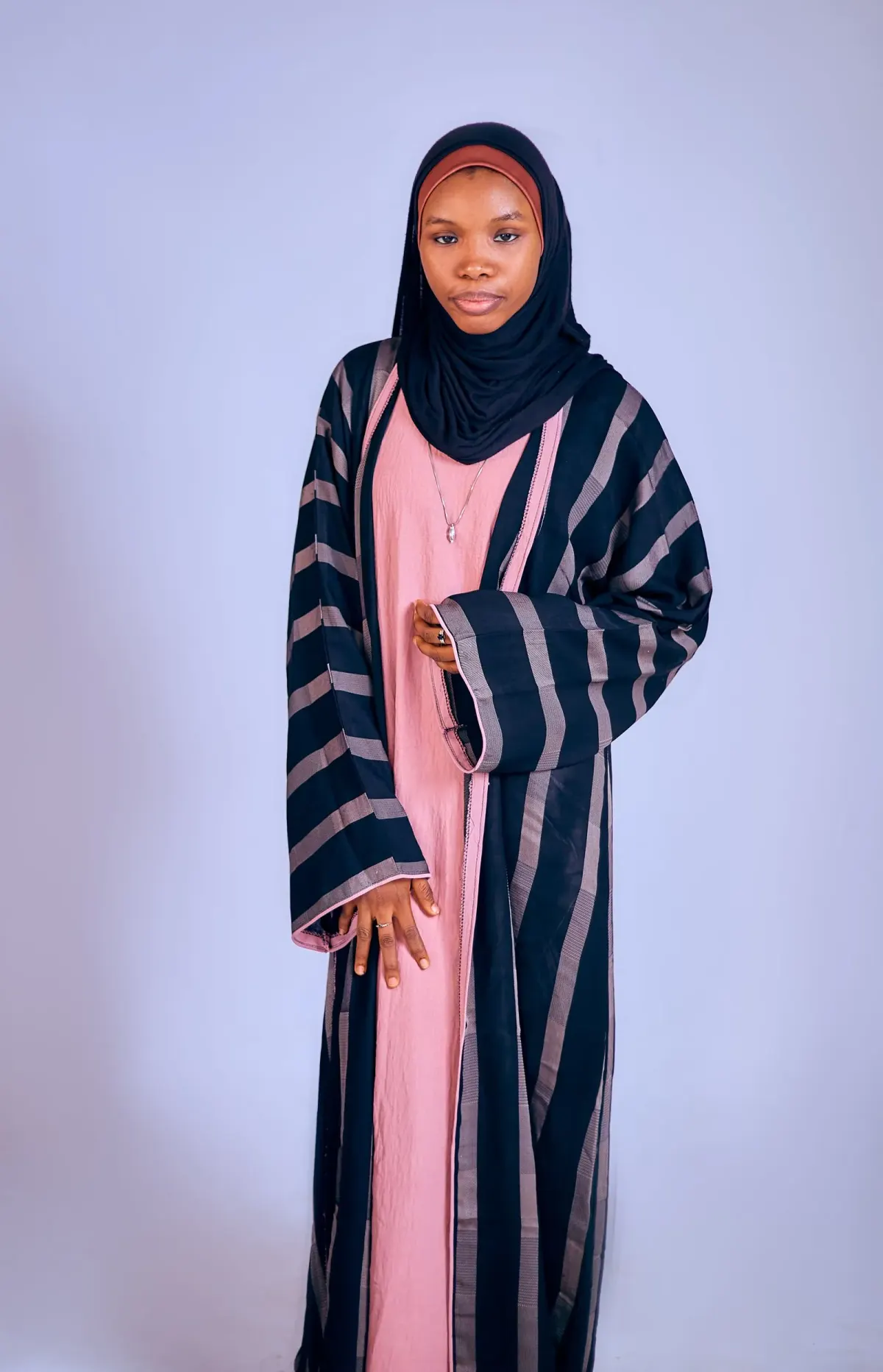 Perempuan berhijab hitam memakai abaya depan terbuka bergaris hitam dan mauve dengan lengan lebar, dipakai di atas gamis pink polos, berlatar studio lavender.