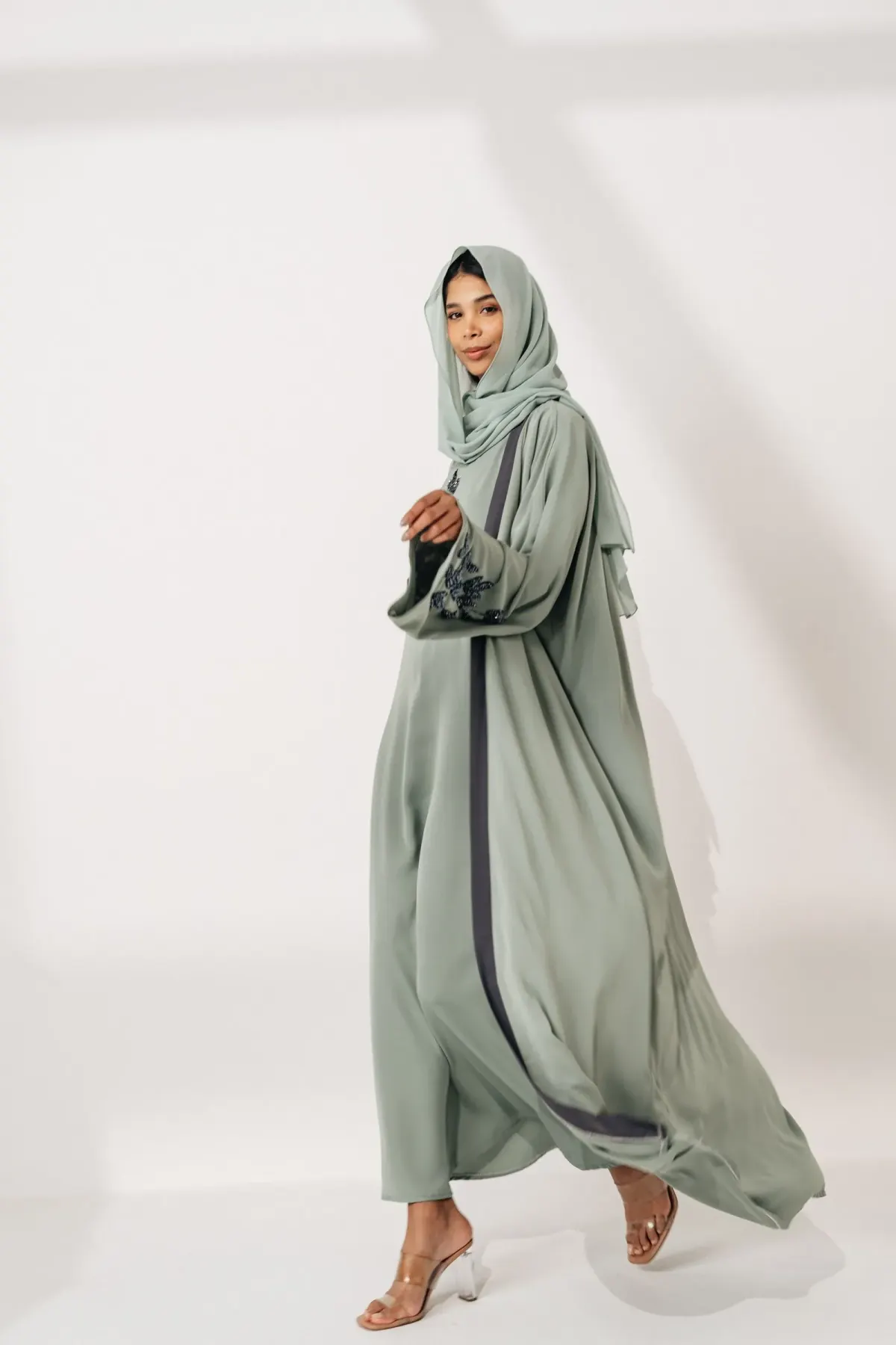 Perempuan berjilbab sage melangkah memakai abaya panjang sage berlengan lebar dengan list abu-abu tua di bagian depan dan bordir di lengan, serta sandal hak bening, di studio berlatar putih.