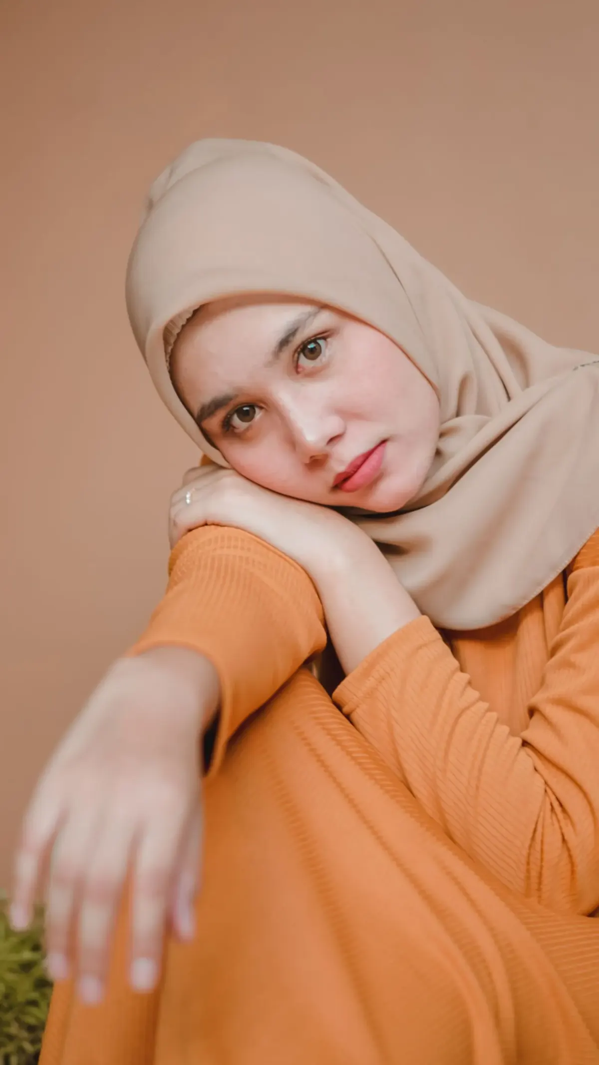 Perempuan berhijab segi empat beige memakai busana rajut bergaris halus berwarna oranye kecokelatan, duduk bertopang lutut di depan dinding cokelat muda.