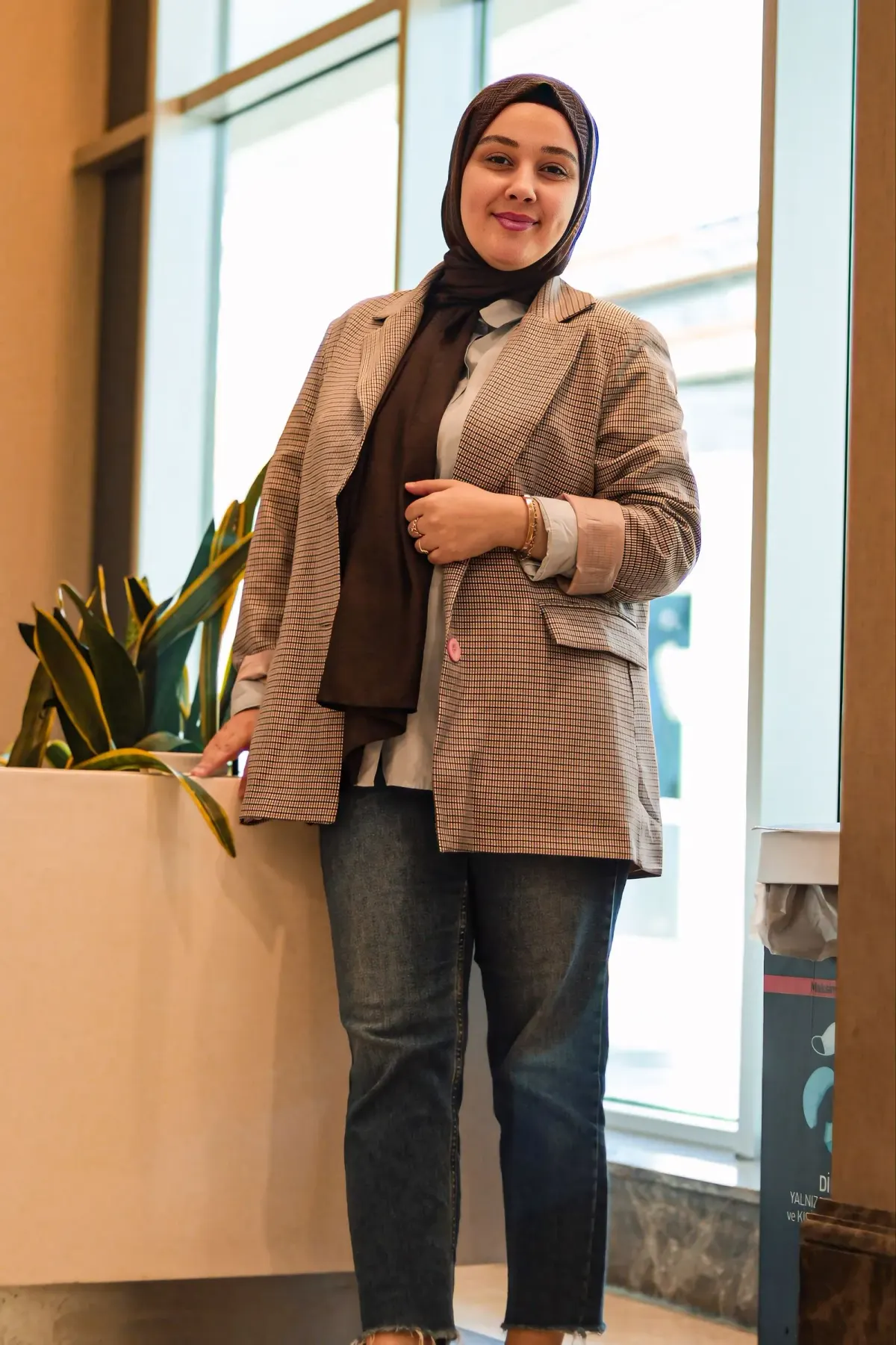 Perempuan berhijab pashmina cokelat tua memakai blazer kotak kecil cokelat dengan lengan digulung, kemeja abu-abu muda, dan jins gelap berujung tak dijahit, berdiri di lobi berjendela besar.