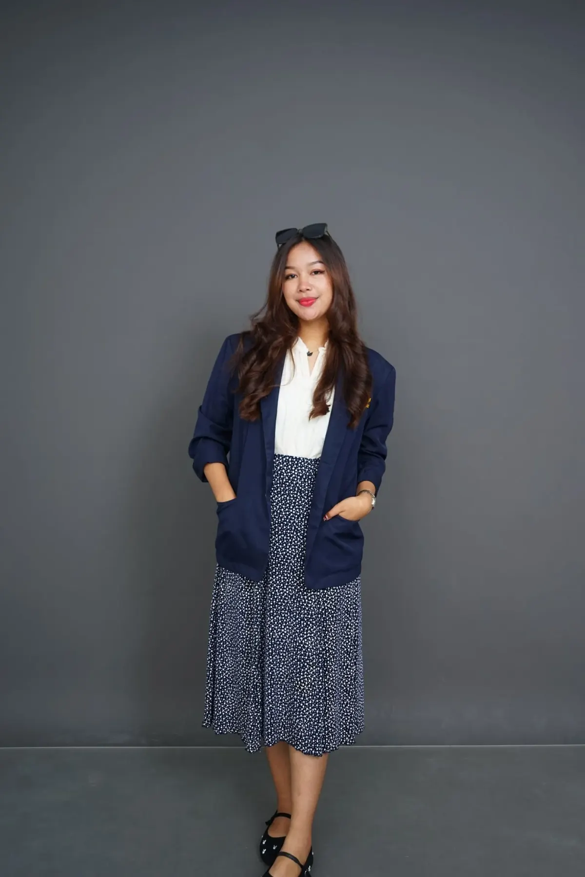 Perempuan berambut panjang memakai blazer navy dengan lengan digulung, blus putih, rok midi navy bermotif bintik putih, dan sepatu hitam bertali, berdiri di depan dinding abu-abu.