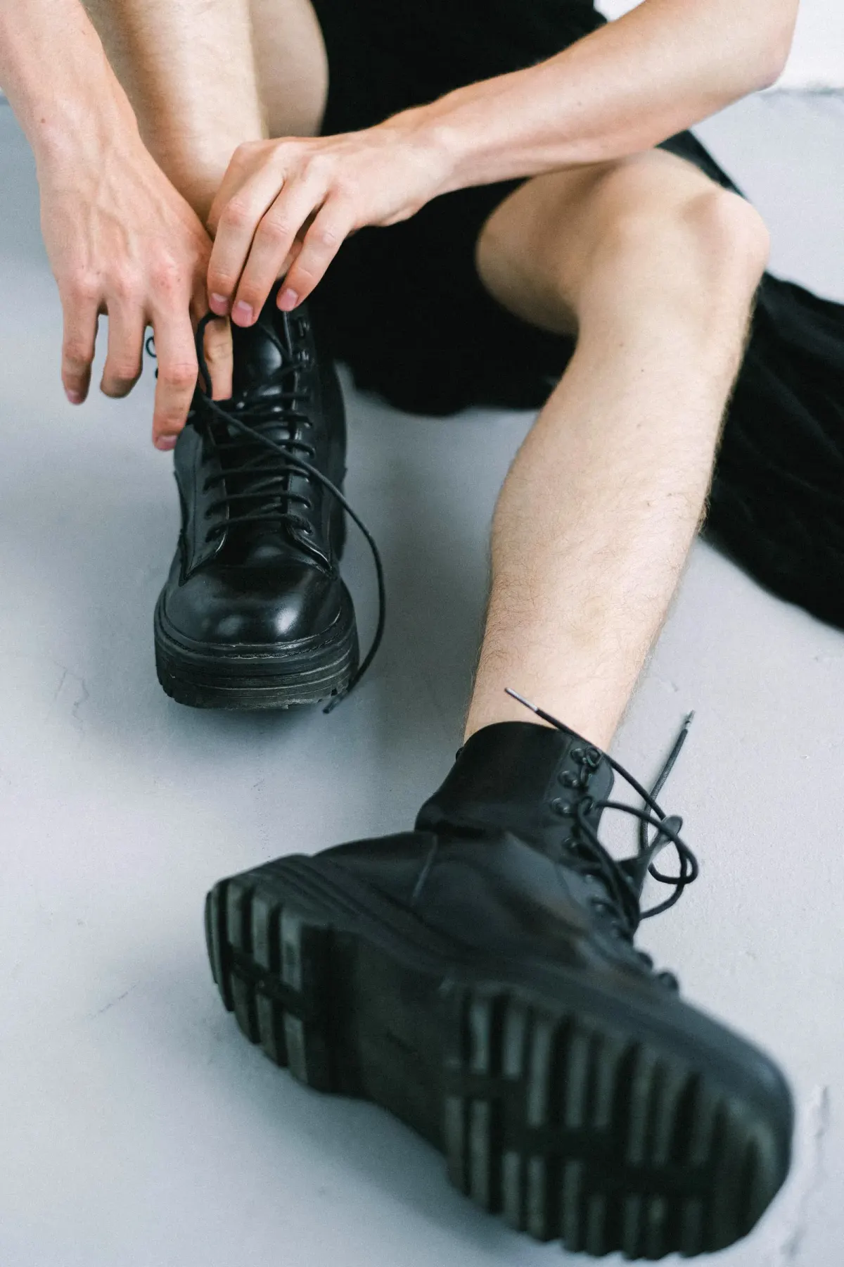 Sepasang boots hitam mengilap bertali dengan sol tebal bergerigi dipakai seseorang yang duduk di lantai putih sambil mengikat tali, wajah tidak terlihat.