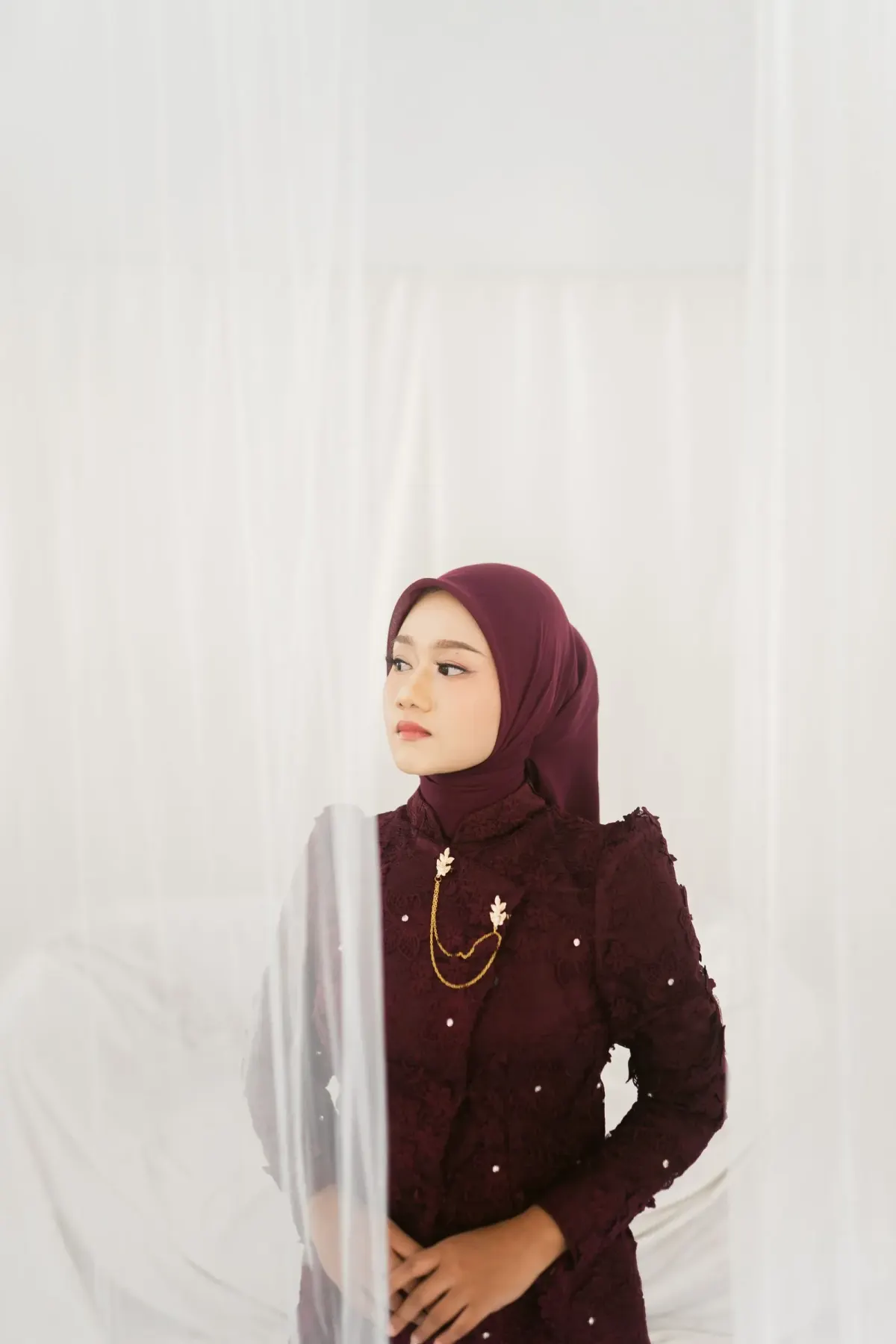 Perempuan berjilbab segi empat maroon dan busana lengan panjang maroon bertekstur dengan aksen mutiara kecil dan bros rantai emas, berdiri di balik tirai putih tipis.