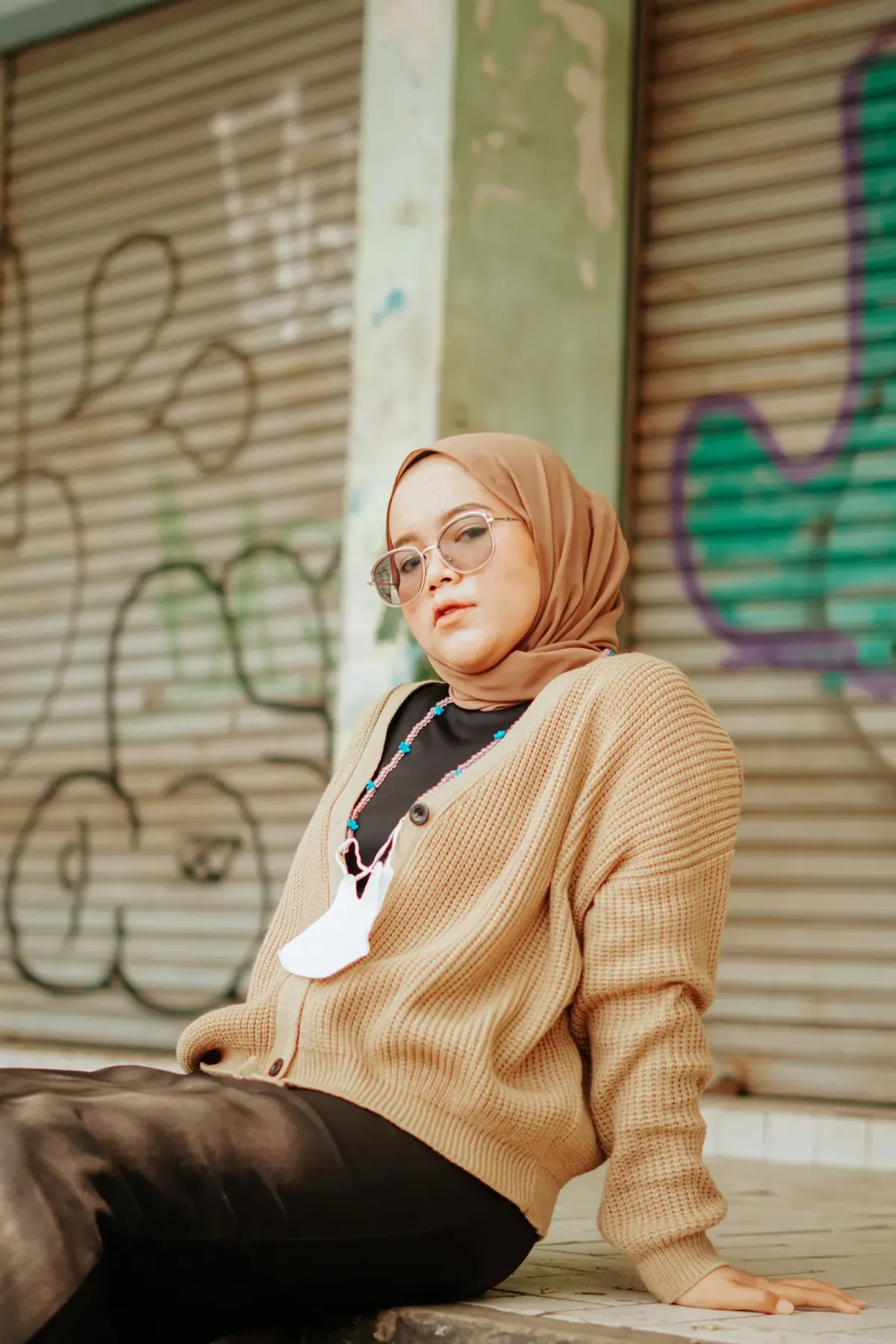 Perempuan berhijab cokelat susu dan berkacamata memakai cardigan rajut cokelat susu berkancing di atas atasan hitam dan celana panjang hitam, duduk di trotoar depan rolling door bergrafiti.