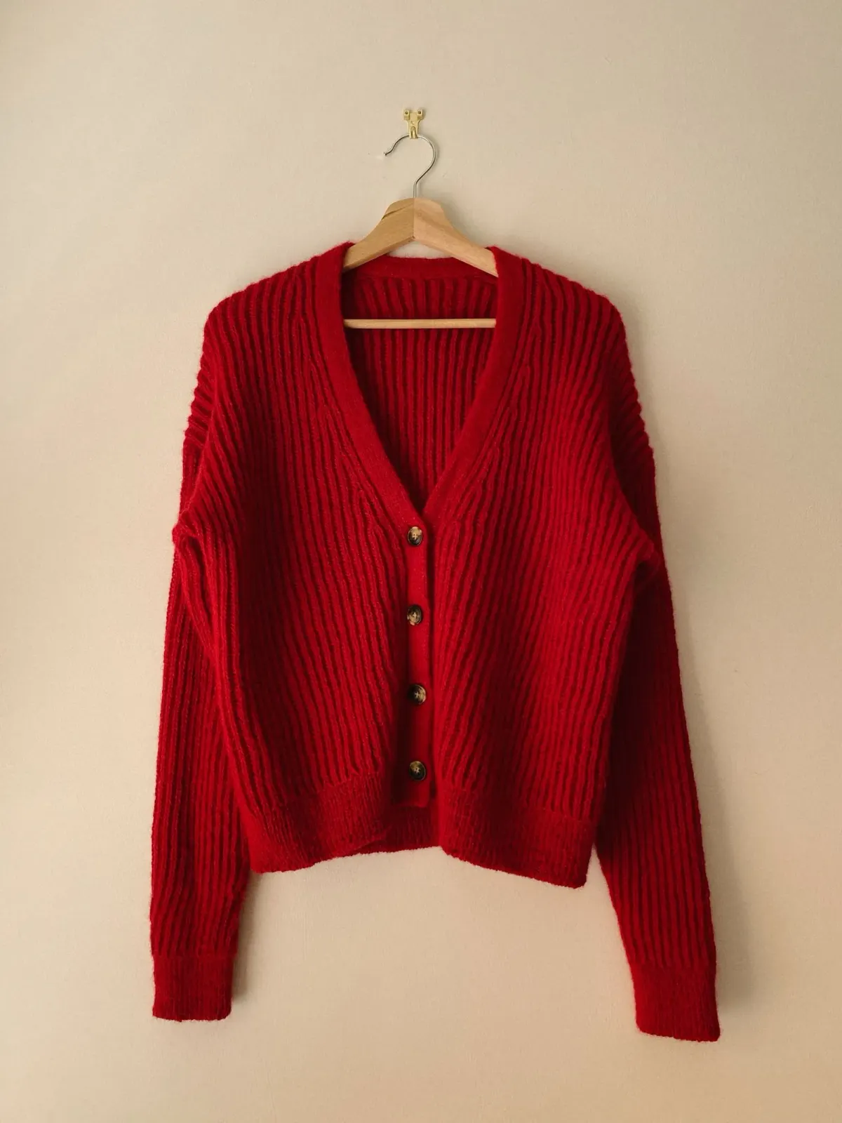 Cardigan rajut merah berleher V dengan empat kancing cokelat, rajutan bergaris vertikal, dan ujung lengan serta keliman rib, digantung di gantungan kayu pada dinding krem.