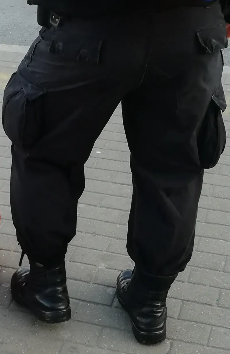Tampak belakang orang memakai celana cargo hitam longgar dengan saku besar berpenutup di sisi kedua paha, ujung celana dimasukkan ke sepatu bot hitam.