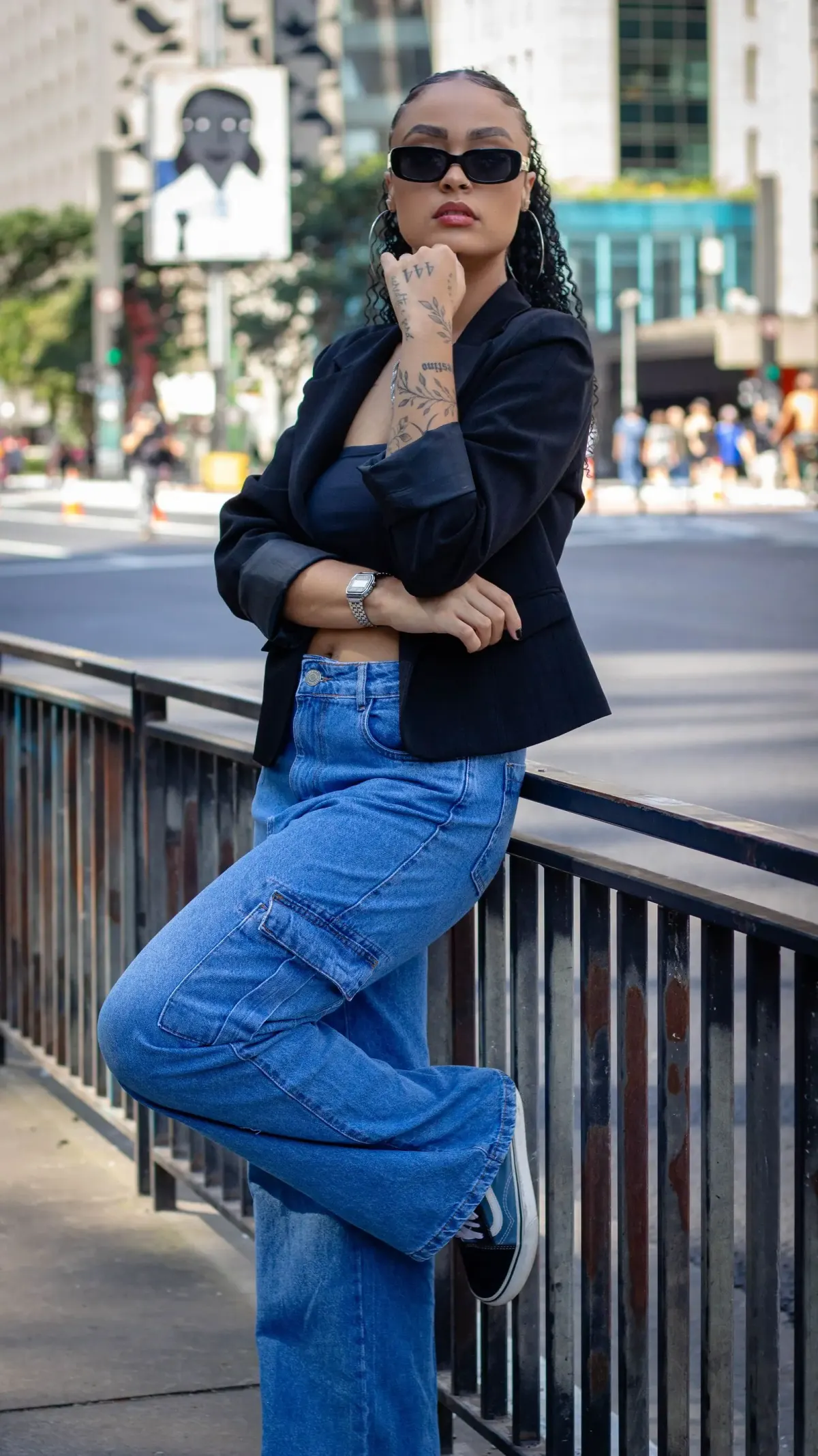 Perempuan berkacamata hitam memakai blazer hitam di atas atasan hitam, celana cargo denim biru berpotongan lebar, dan sepatu kets hitam, bersandar di pagar besi di pinggir jalan kota.