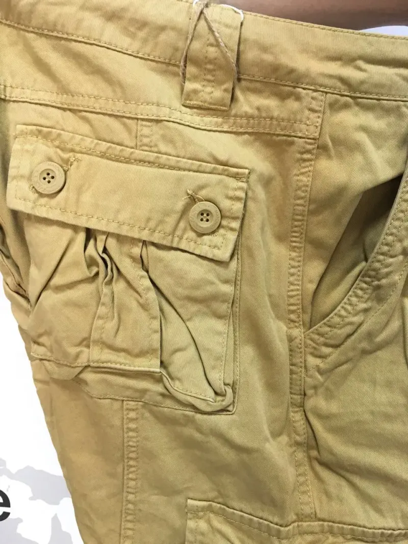 Close-up saku cargo pada celana khaki, saku tempel berlipat di sisinya dengan penutup berkancing dua dan jahitan ganda di sekelilingnya.