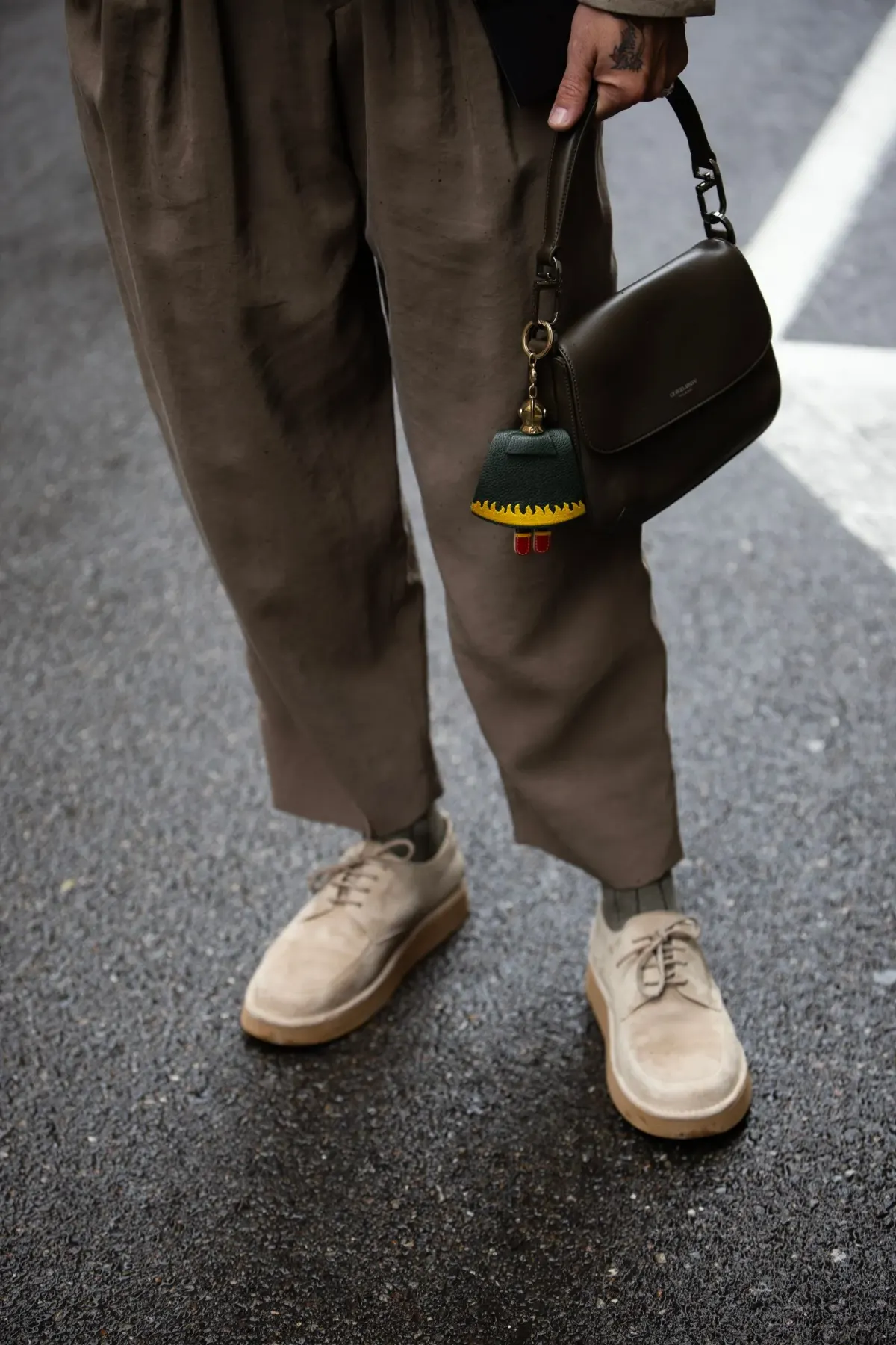 Tampilan pinggang ke bawah seseorang memakai celana cokelat berlipit dengan kaki lebar yang berhenti di atas mata kaki, kaus kaki gelap, dan sepatu suede krem bertali, sambil menjinjing tas hitam di jalan beraspal.