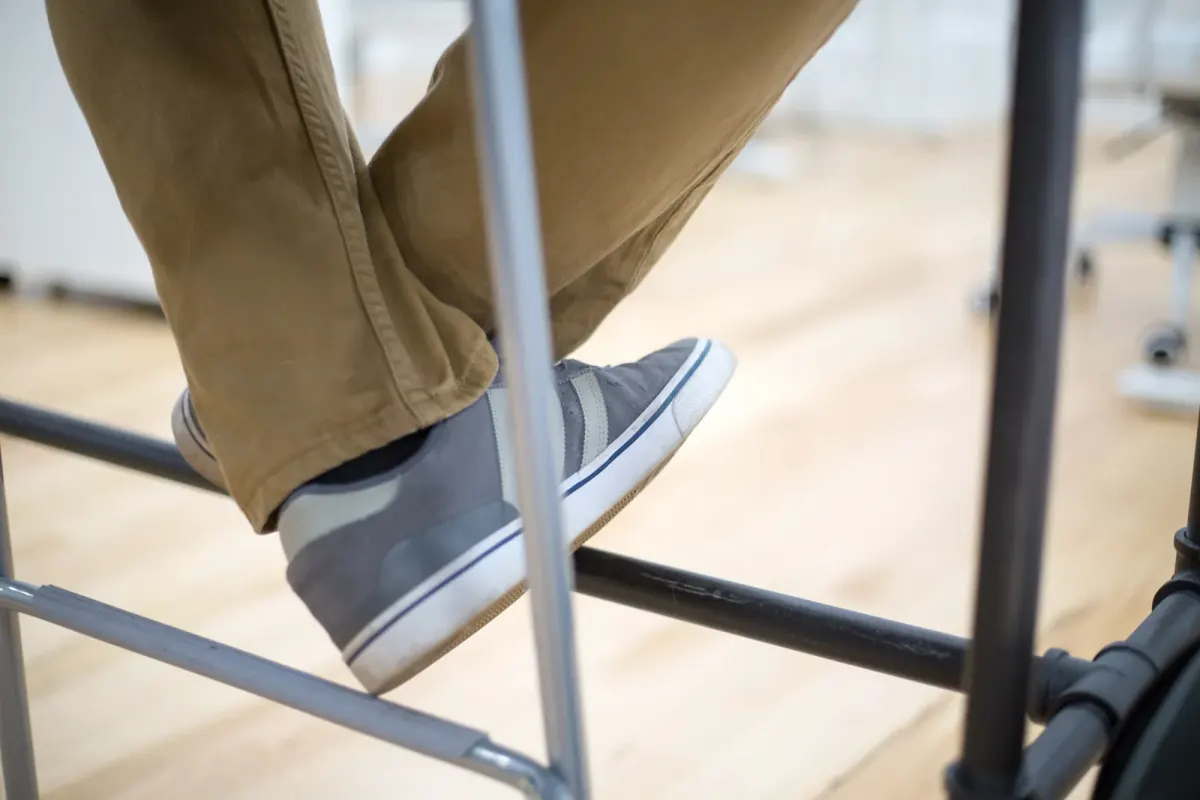 Detail kaki pria memakai celana chino khaki dan sneakers kanvas abu-abu bersol putih, bertumpu pada kaki kursi logam di ruangan berlantai kayu.