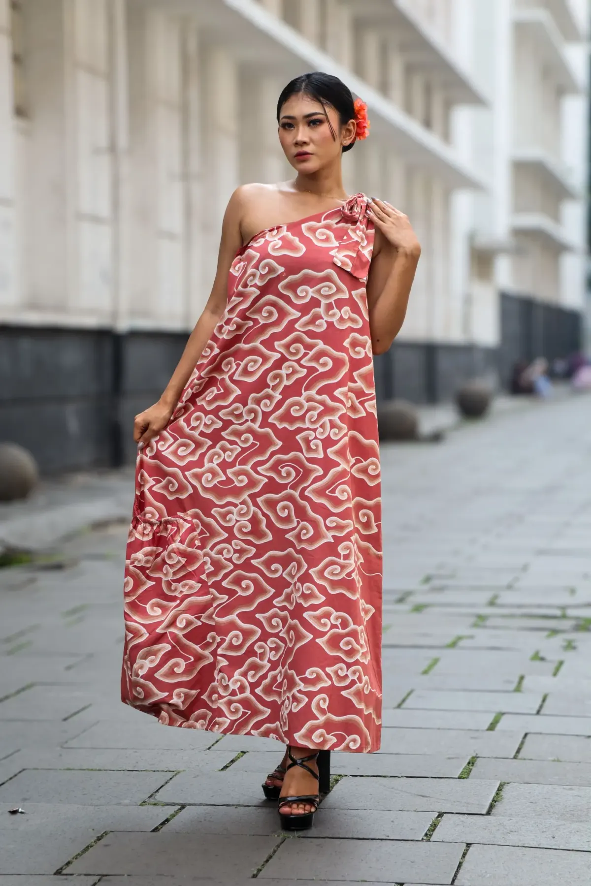 Perempuan berambut disanggul dengan bunga merah memakai dress batik satu bahu warna terakota bermotif awan krem dan sepatu platform hitam, berdiri di jalan berbatu, Jakarta.