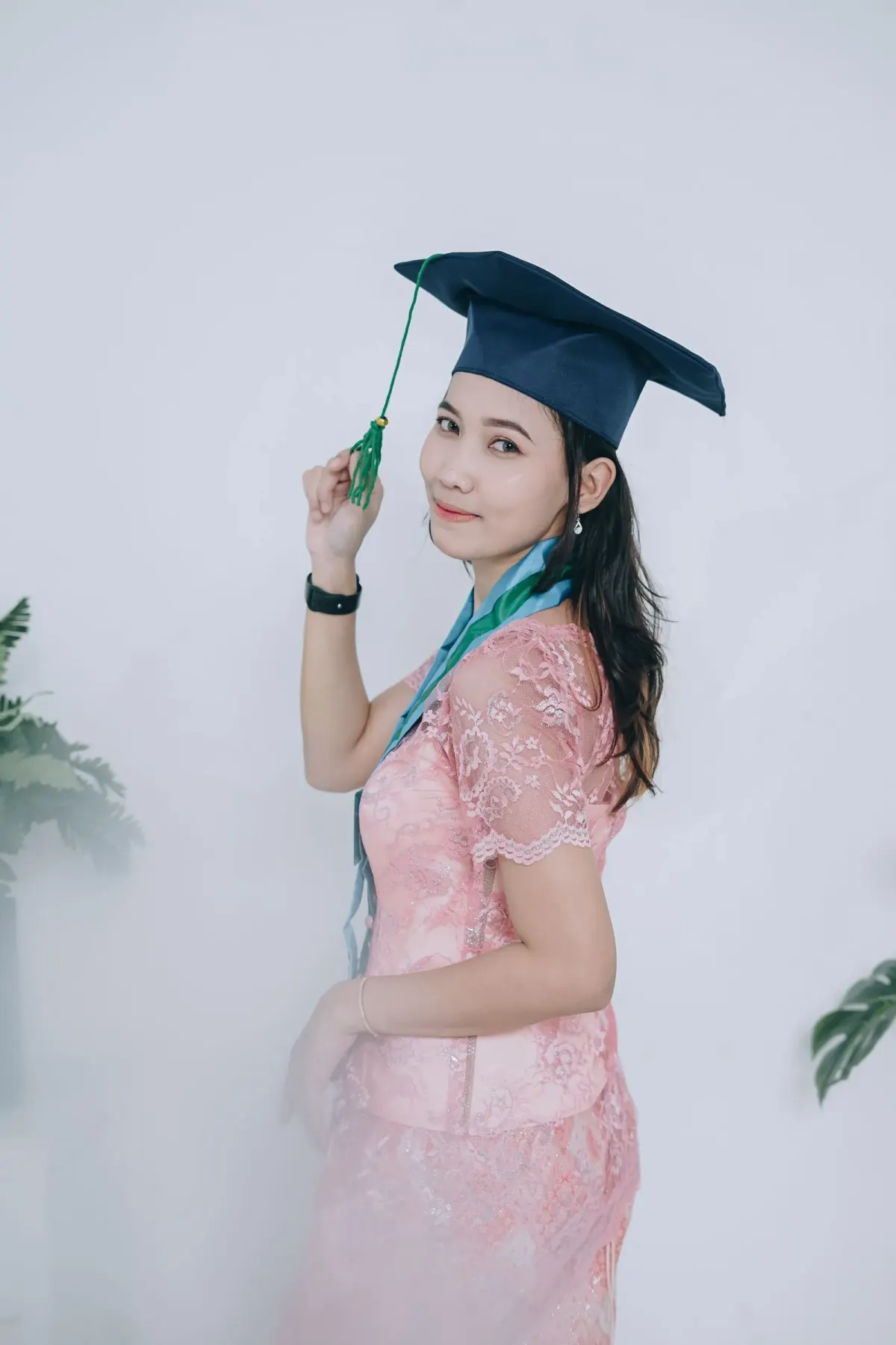 Perempuan berambut panjang memakai dress brokat merah muda berlengan pendek, selempang hijau kebiruan, dan topi toga navy berumbai hijau, berpose di studio putih.