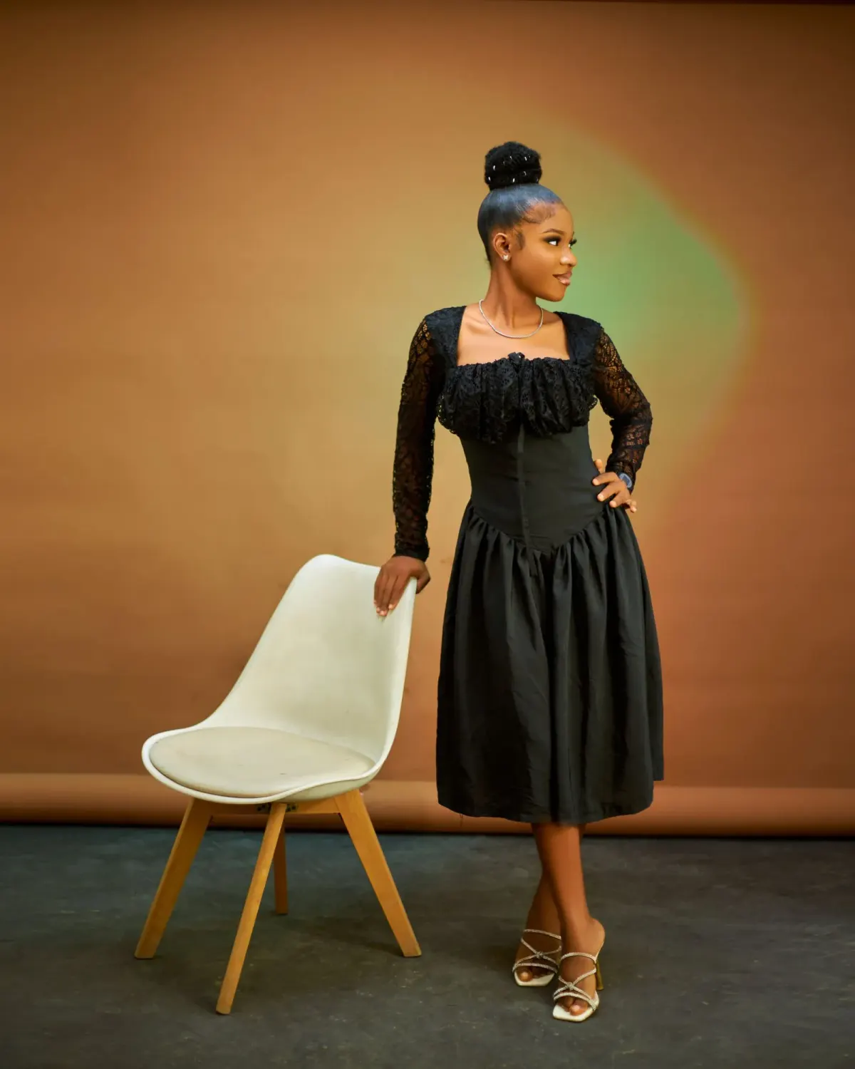 Perempuan memakai dress hitam selutut lebih dengan atasan renda lengan panjang, badan pas, dan rok mengembang, dipadukan sandal hak bertali, berdiri di samping kursi putih di studio.