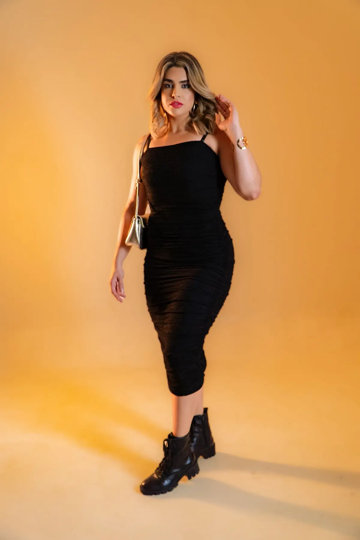 Perempuan memakai dress hitam bertali spageti yang pas badan sampai di bawah lutut, dipadukan bot hitam bertali dan tas bahu kecil berwarna perak, di studio berlatar jingga.