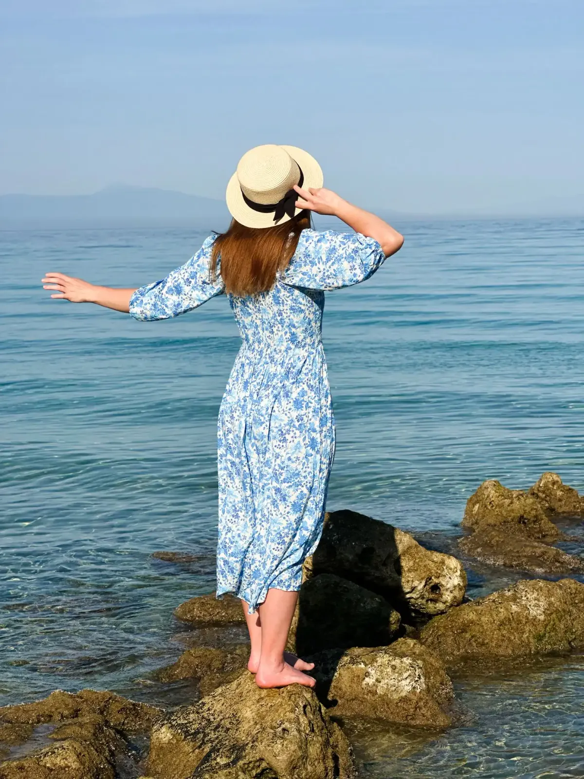 Perempuan dilihat dari belakang memakai dress midi putih bermotif bunga biru dengan lengan mengembang dan topi jerami berpita hitam, berdiri tanpa alas kaki di batu tepi laut.