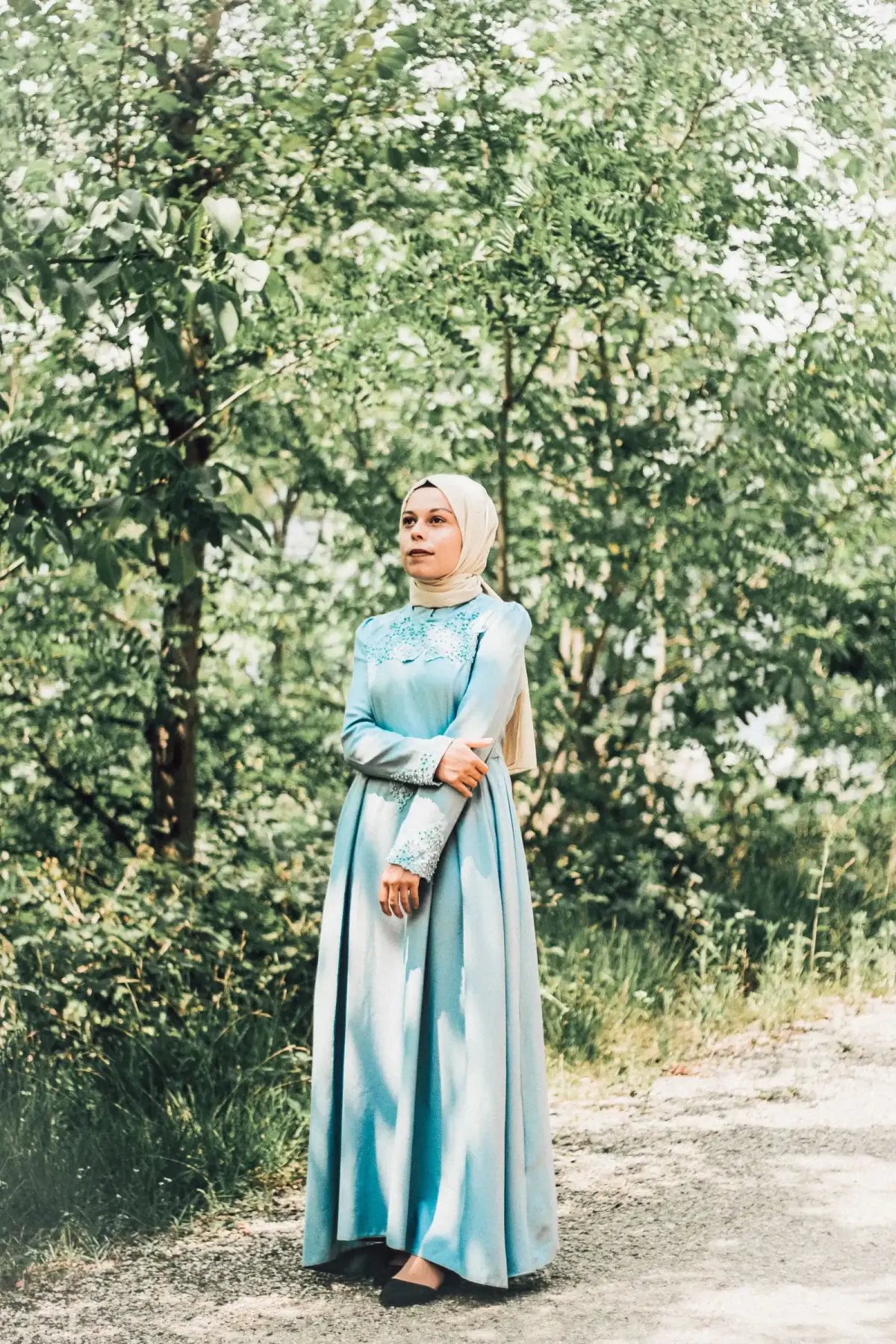 Perempuan berhijab krem memakai gamis biru muda panjang dengan brokat di dada dan lengan serta flat shoes hitam, berdiri di jalan setapak di antara pepohonan.