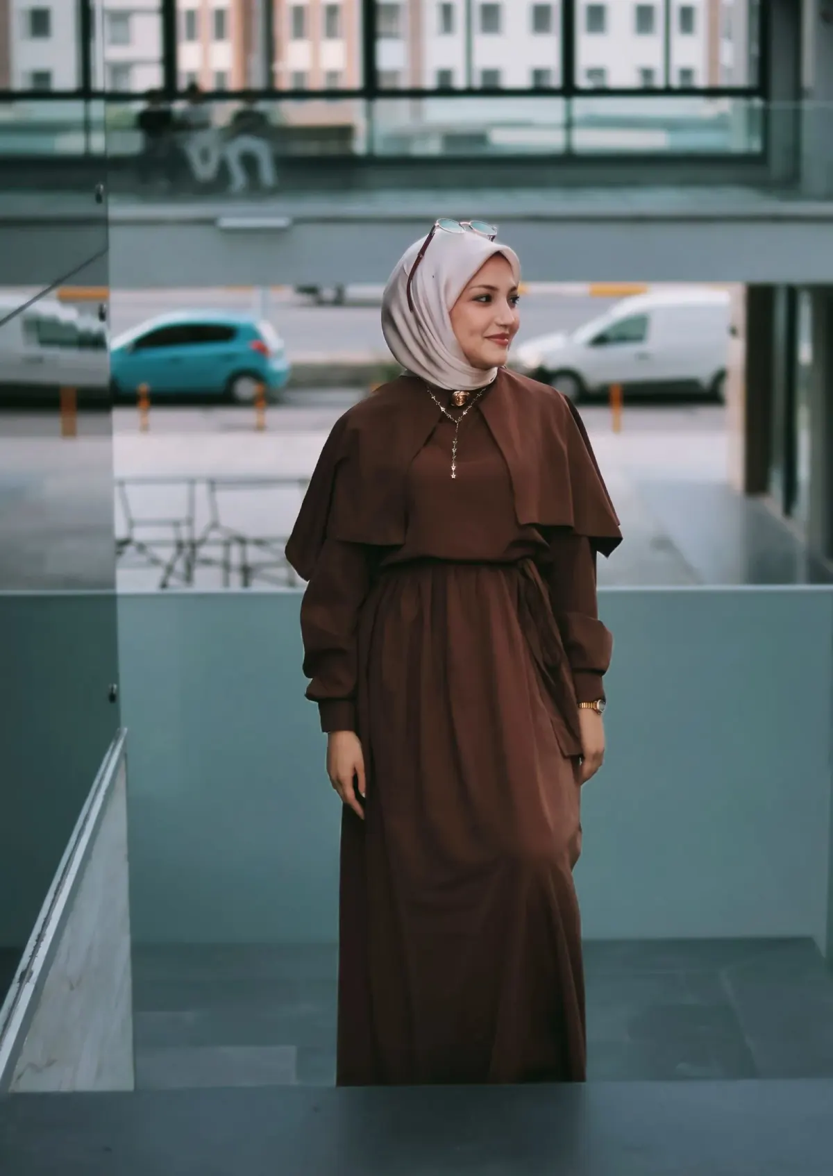 Perempuan berhijab merah muda keabuan memakai gamis cokelat tua berlapis cape dengan tali pinggang dan kalung, berdiri di area gedung berkaca.