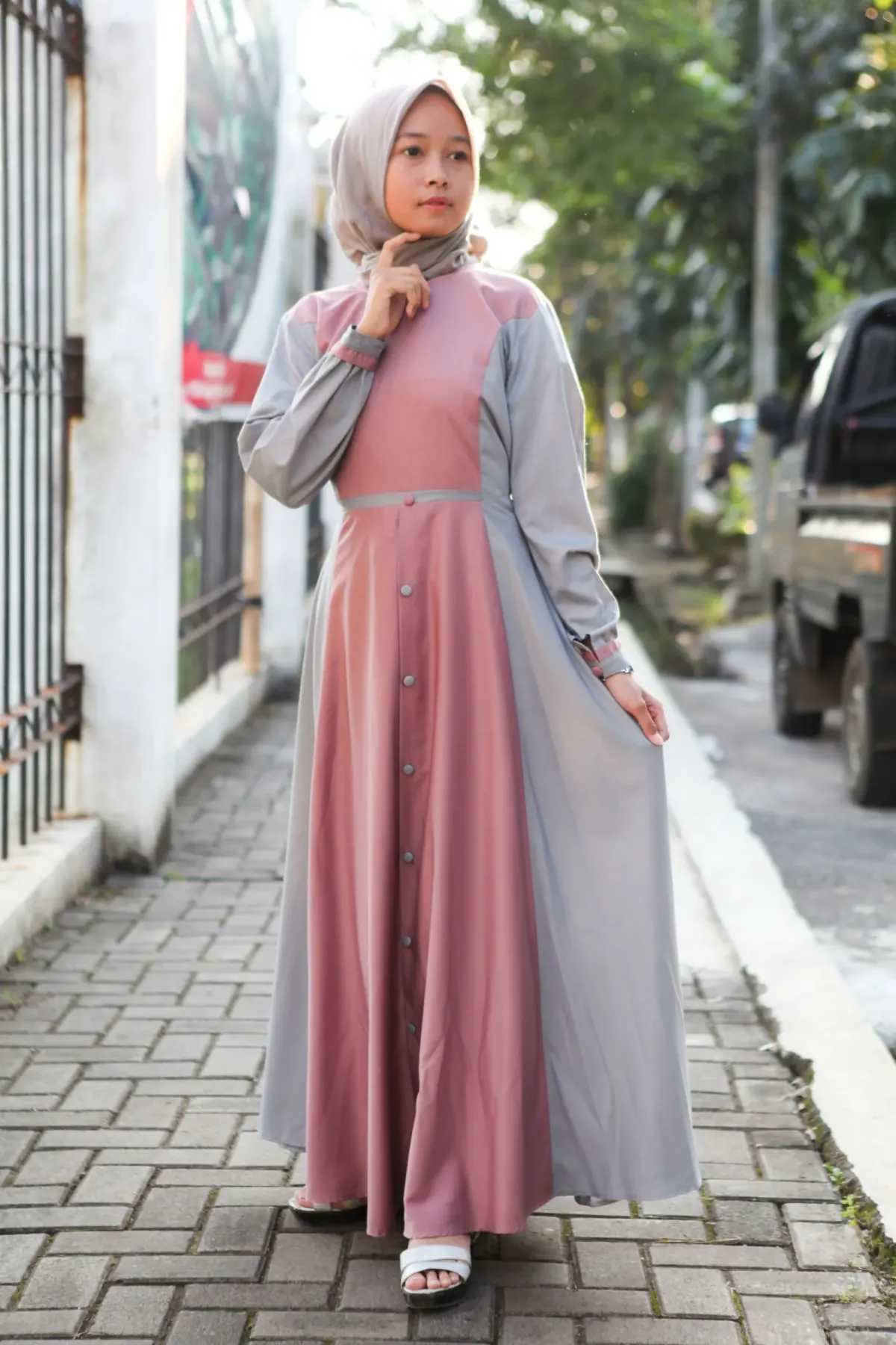 Perempuan berhijab krem memakai gamis kombinasi dua warna: panel depan pink salem berkancing dari pinggang sampai bawah, panel samping dan lengan abu-abu, sabuk tipis abu di pinggang, dan sandal putih.