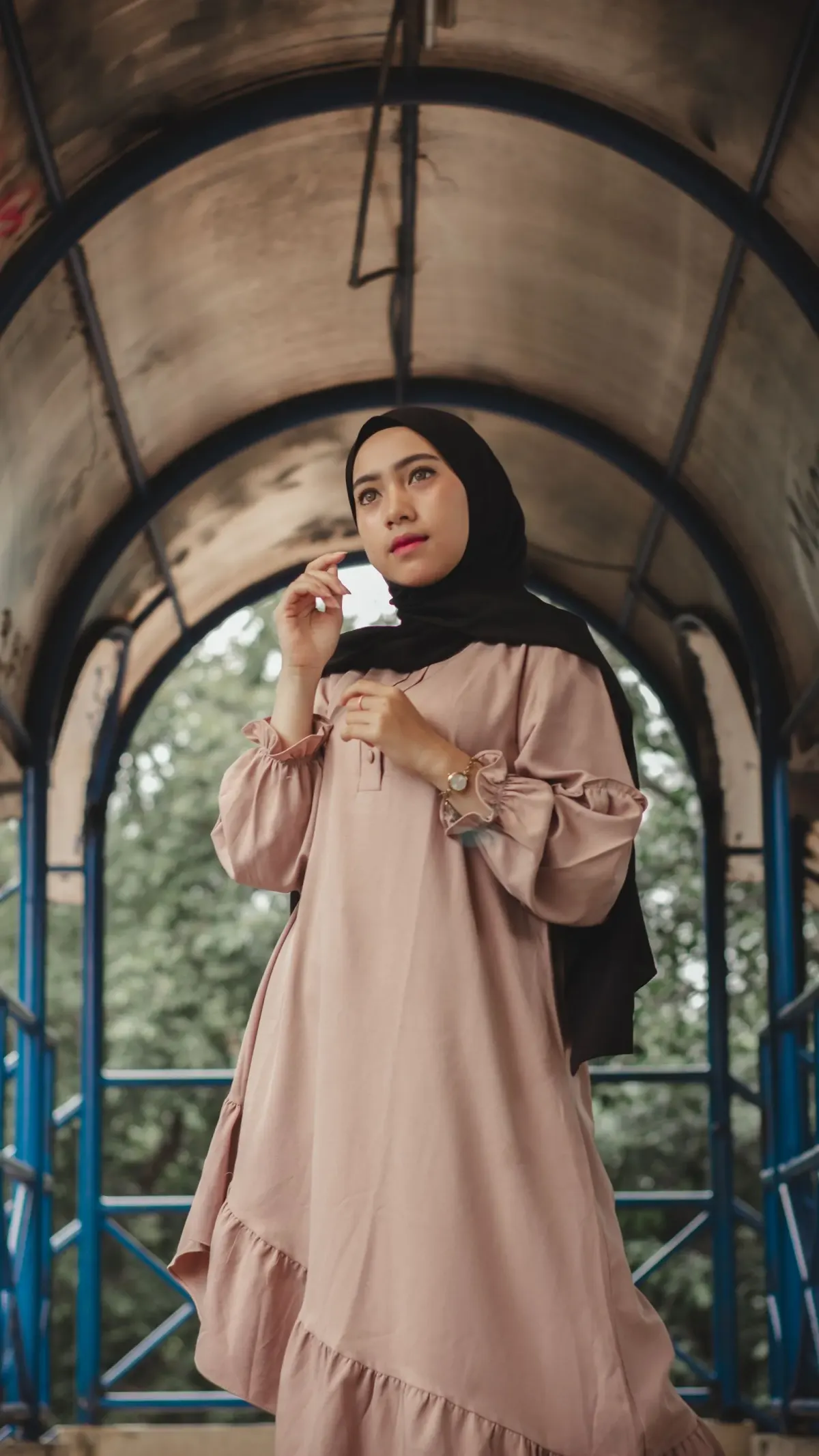 Perempuan berhijab hitam memakai gamis krem longgar dengan lengan balon bermanset kerut dan ruffle miring di bagian bawah, berdiri di lorong beratap lengkung.