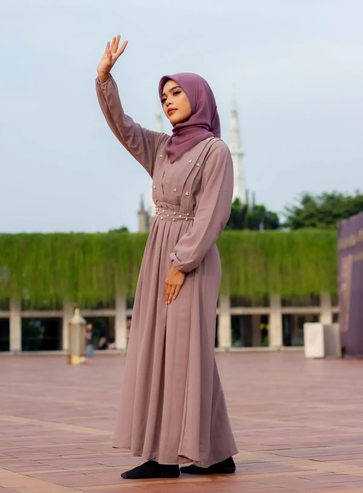Perempuan berhijab ungu tua memakai gamis merah muda keabuan berhias mutiara dengan pinggang berlipit, mengangkat satu tangan, berdiri di pelataran berlantai bata.