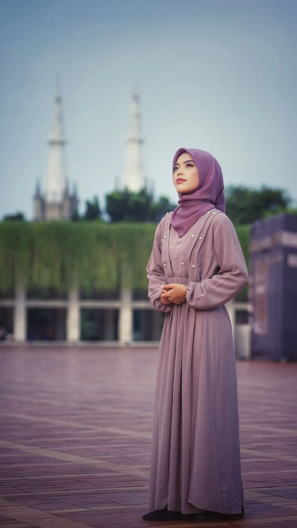 Perempuan berhijab ungu memakai gamis mauve lengan panjang bermanset dengan pinggang berkerut dan detail mutiara kecil di dada, berdiri di pelataran dengan dua menara di latar belakang.