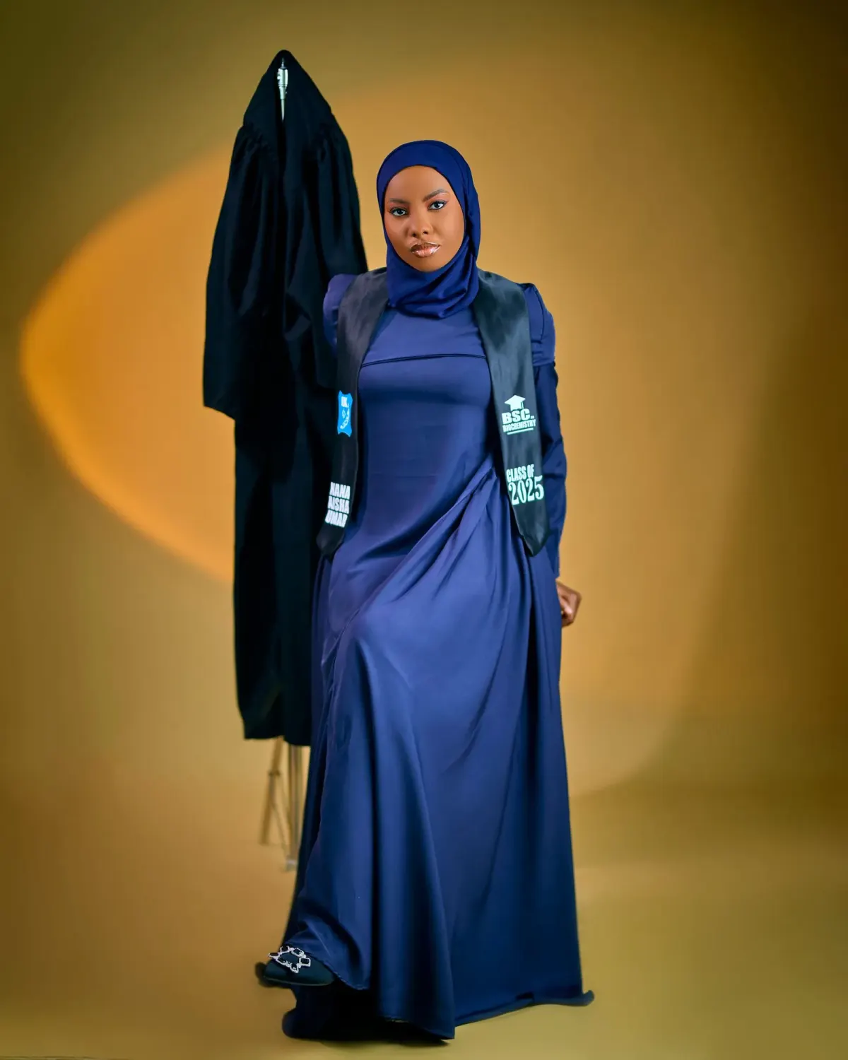 Perempuan berjilbab navy memakai gamis navy panjang yang tampak mengilap dan selempang wisuda hitam bertulisan angkatan, berdiri di samping toga hitam yang digantung, latar studio kuning.