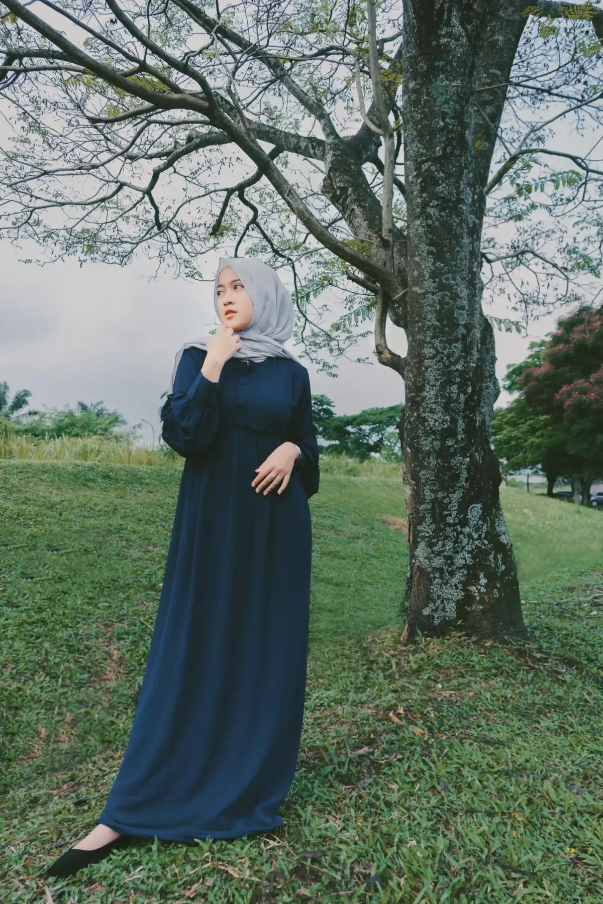 Perempuan berhijab abu-abu muda memakai gamis navy polos lengan panjang dan flat shoes hitam, berdiri di padang rumput di bawah pohon besar.