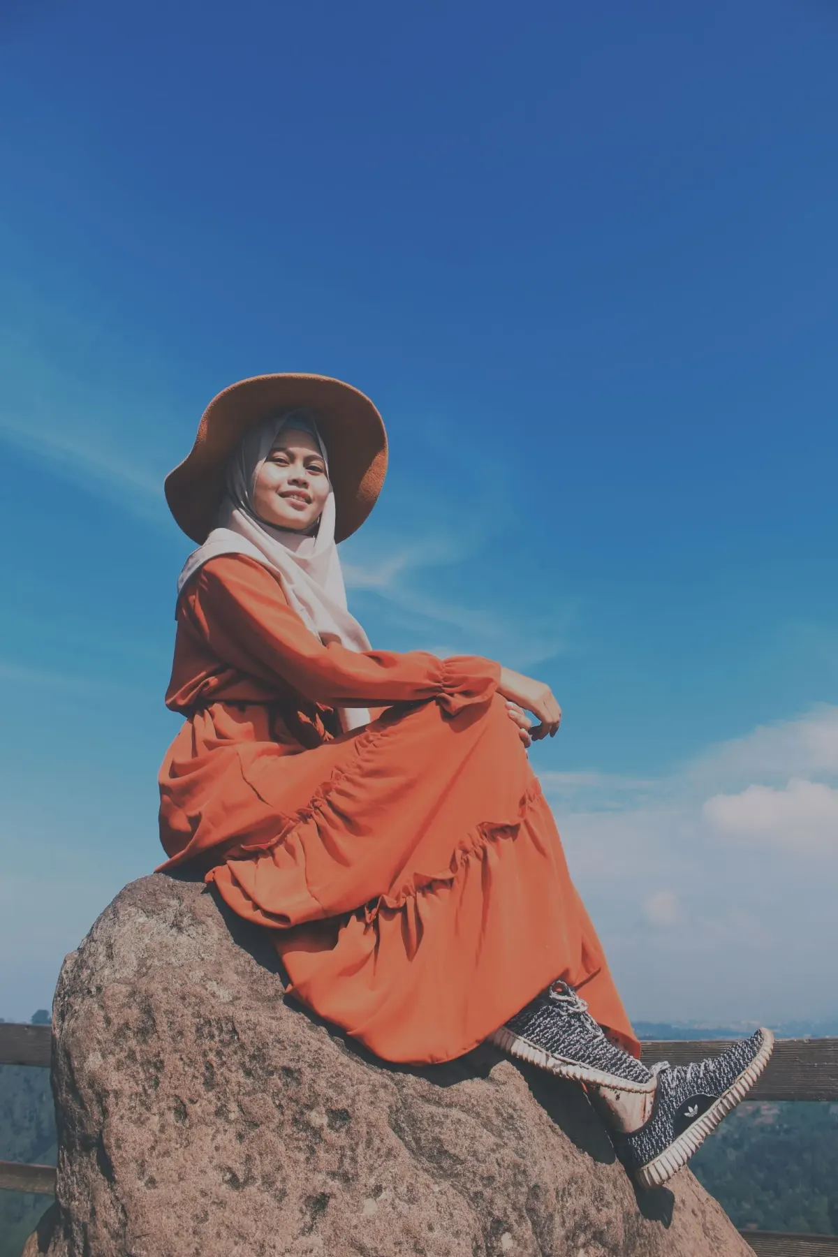 Perempuan berhijab putih memakai gamis oranye bertumpuk, topi lebar cokelat, dan sneakers rajut abu-abu, duduk di atas batu besar dengan latar langit biru.
