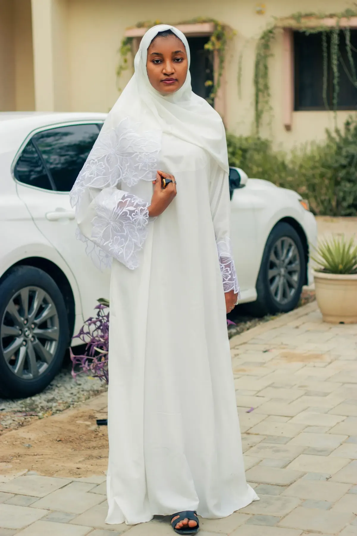 Perempuan memakai abaya putih panjang dengan lengan lebar berbordir renda dan jilbab putih panjang, berdiri di halaman berpaving di depan mobil putih.
