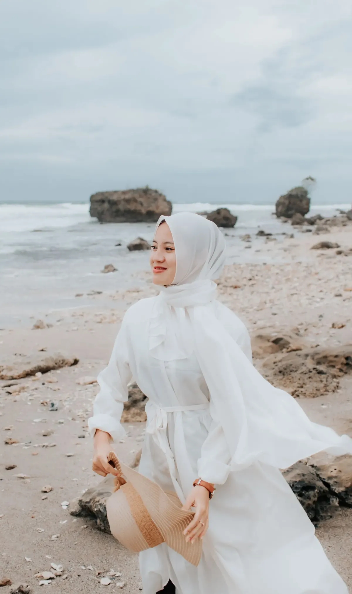 Perempuan berhijab putih memakai gamis putih panjang bertali pinggang dan jam tangan, memegang topi jerami, berjalan di pantai berbatu karang.