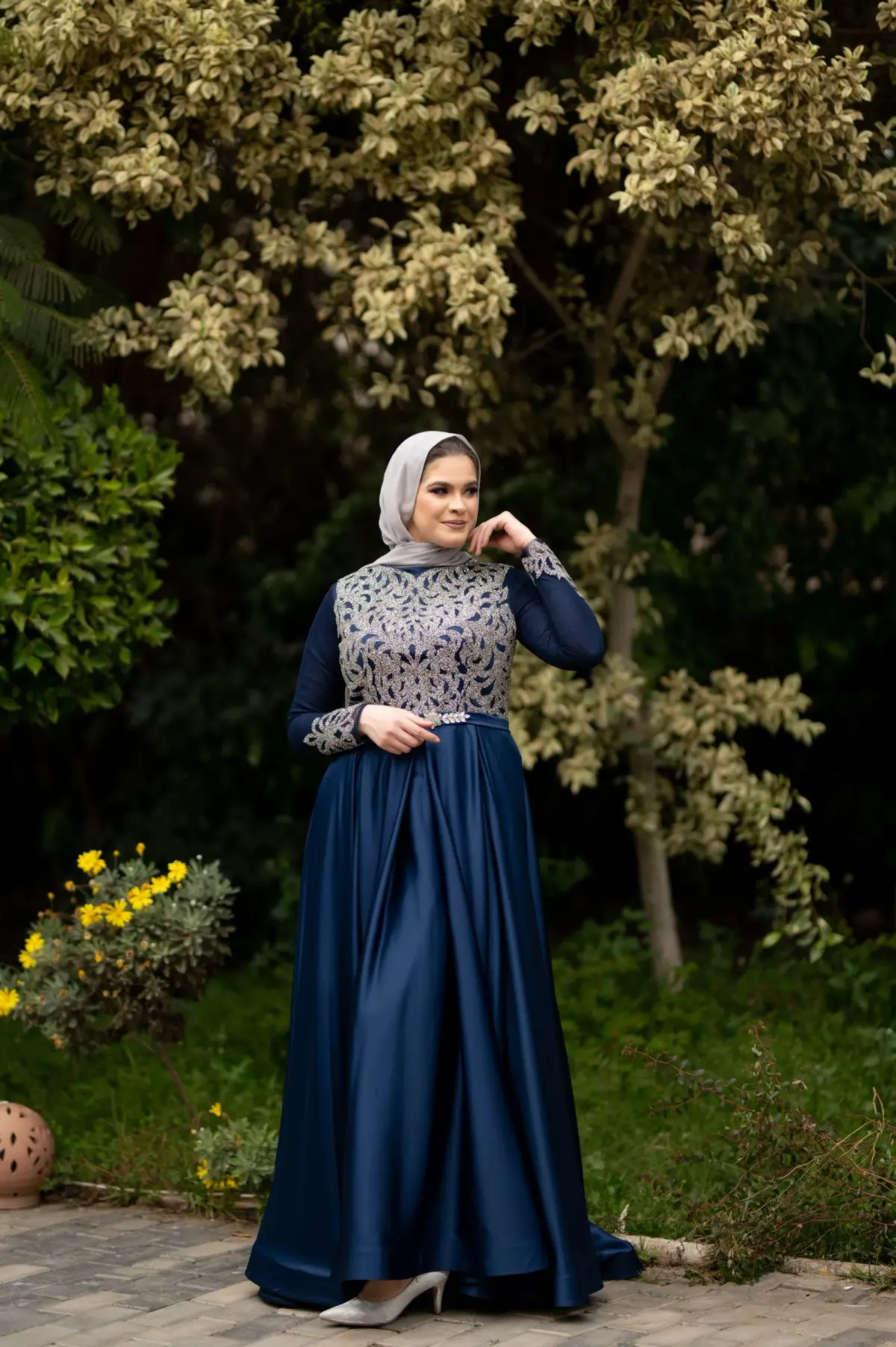Perempuan berhijab abu-abu memakai gaun panjang satin navy dengan bordir perak di bagian atas, sepatu hak putih, berdiri di taman.