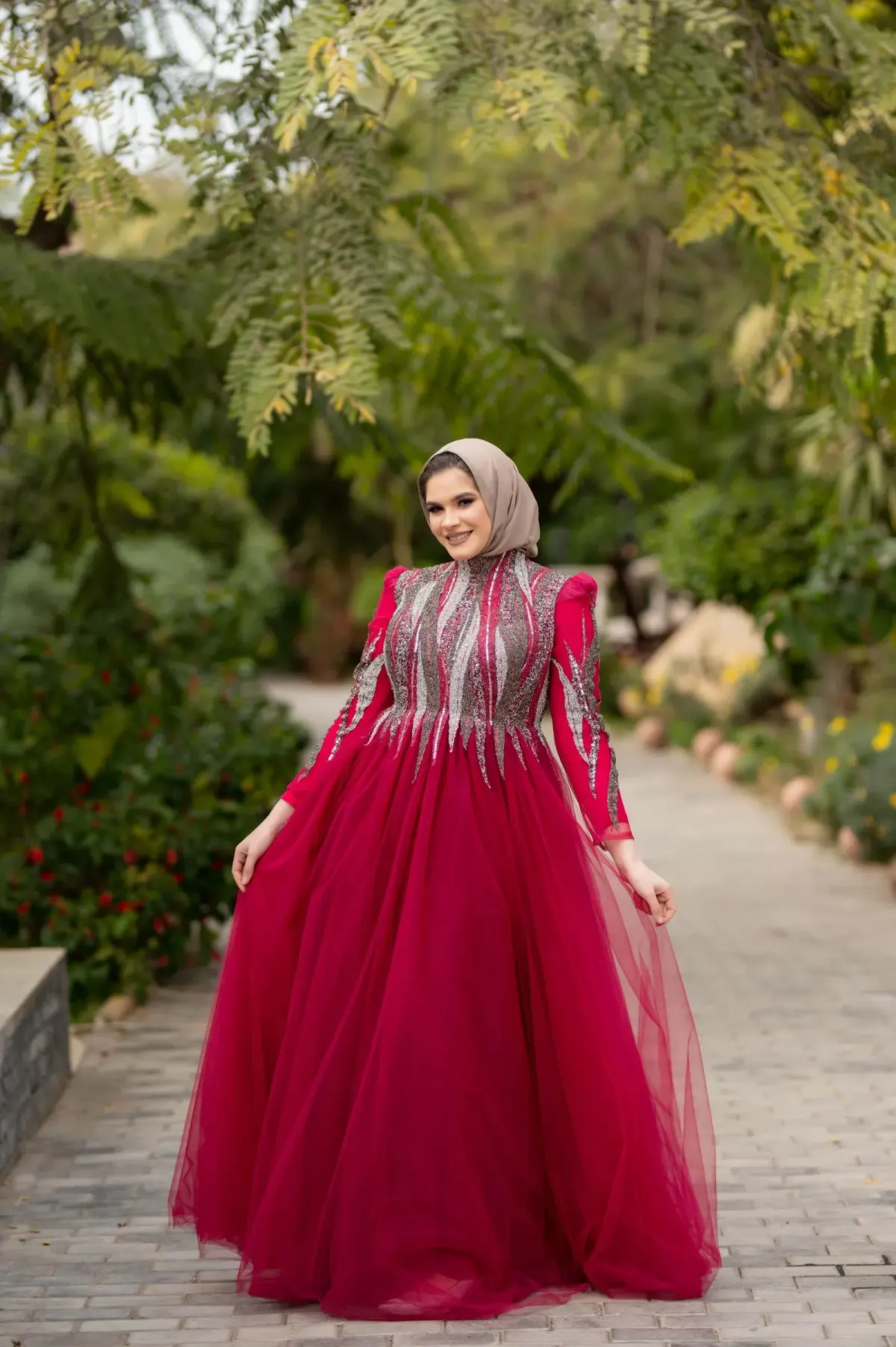 Perempuan berhijab beige memakai gaun pesta merah dengan bordir perak di bagian atas dan rok tule mengembang, berdiri di jalan setapak taman.