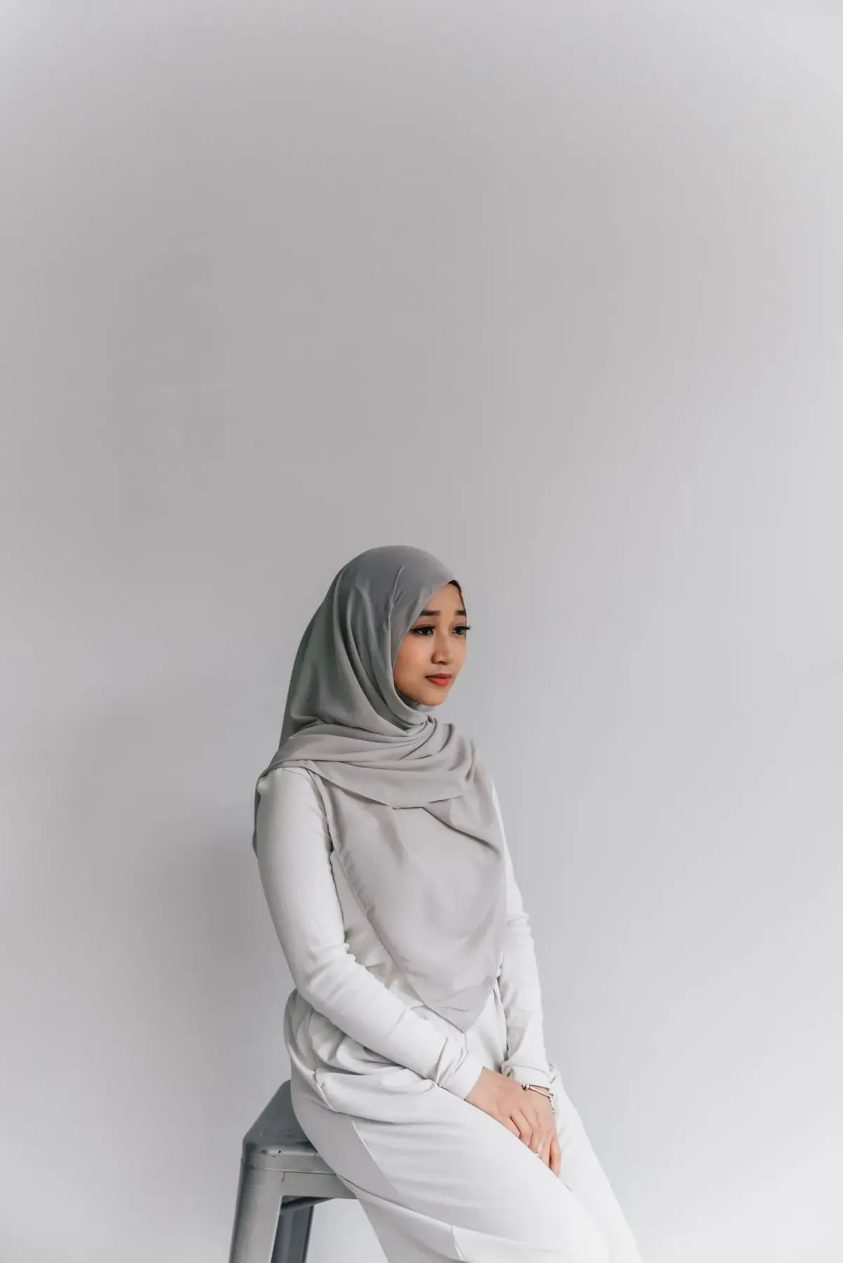 Perempuan duduk di bangku besi di studio berlatar abu-abu, memakai hijab abu-abu tipis yang menjuntai menutupi dada sampai perut, atasan lengan panjang putih, dan celana putih.