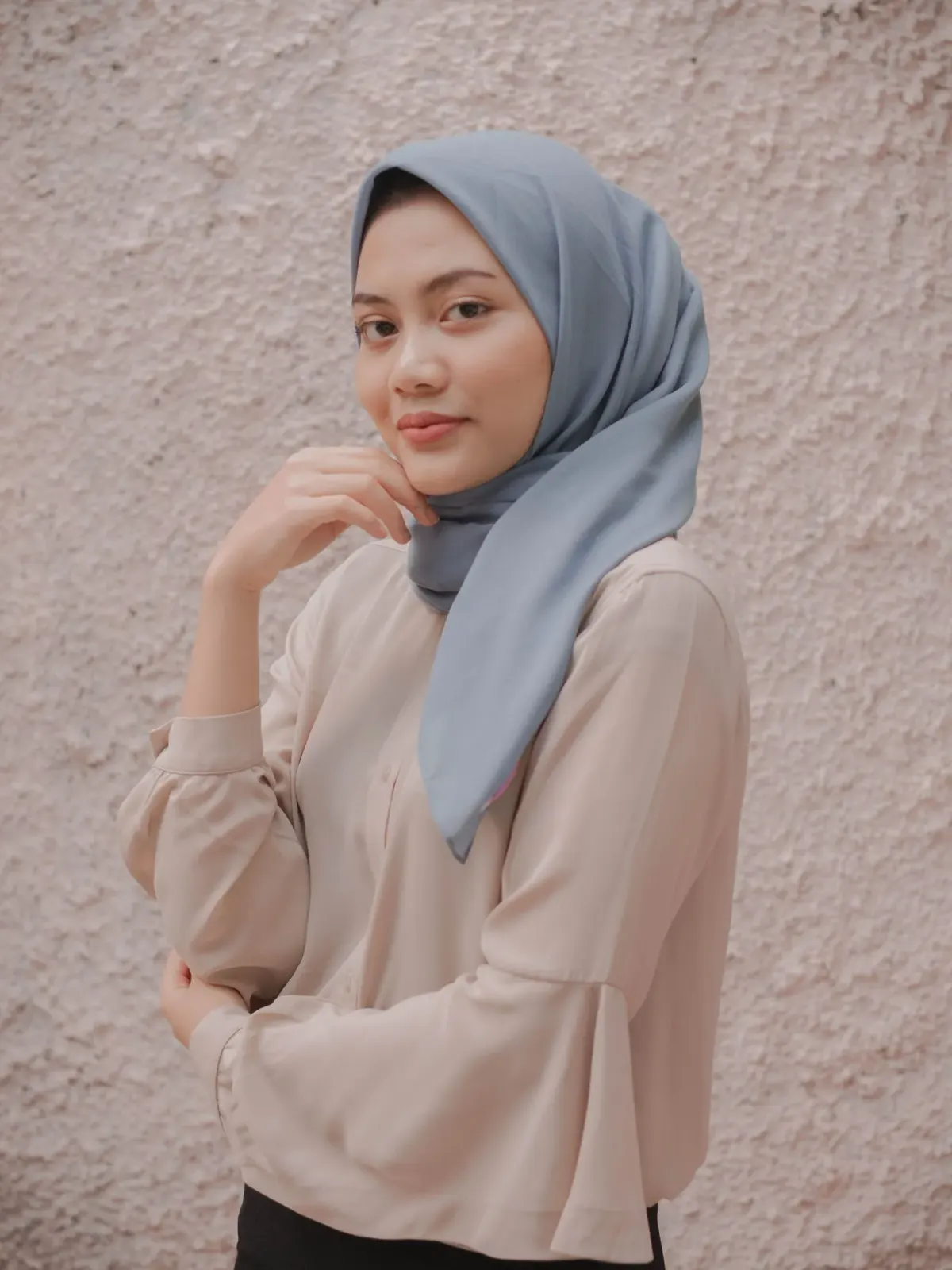 Perempuan berhijab biru keabuan dengan satu ujung kain disampirkan ke bahu kanan, memakai blus krem berlengan lonceng, berpose di depan dinding bertekstur merah muda pucat.