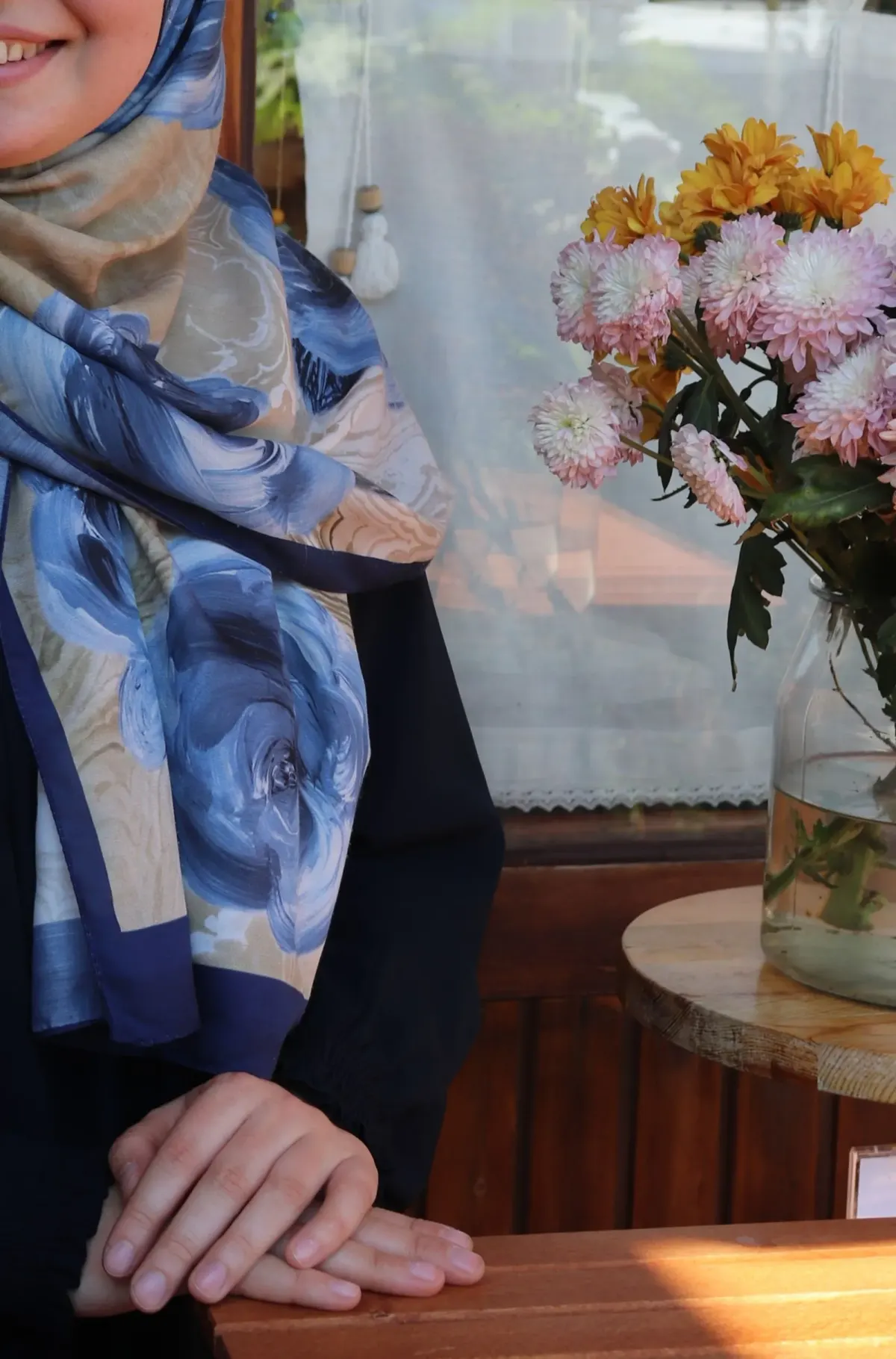 Hijab bermotif lukisan mawar biru di atas latar krem dengan tepi biru tua menjuntai di depan baju hitam, di samping vas bunga krisan merah muda dan kuning di meja kayu.
