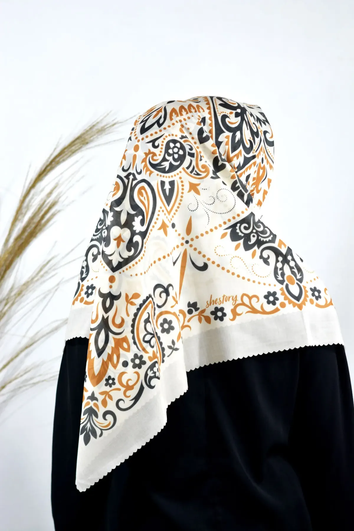 Tampak belakang perempuan berhijab segi empat motif paisley krem, hitam, dan oranye yang dilipat segitiga, sudut kainnya jatuh di punggung, tepi kain bergerigi, di atas baju hitam.