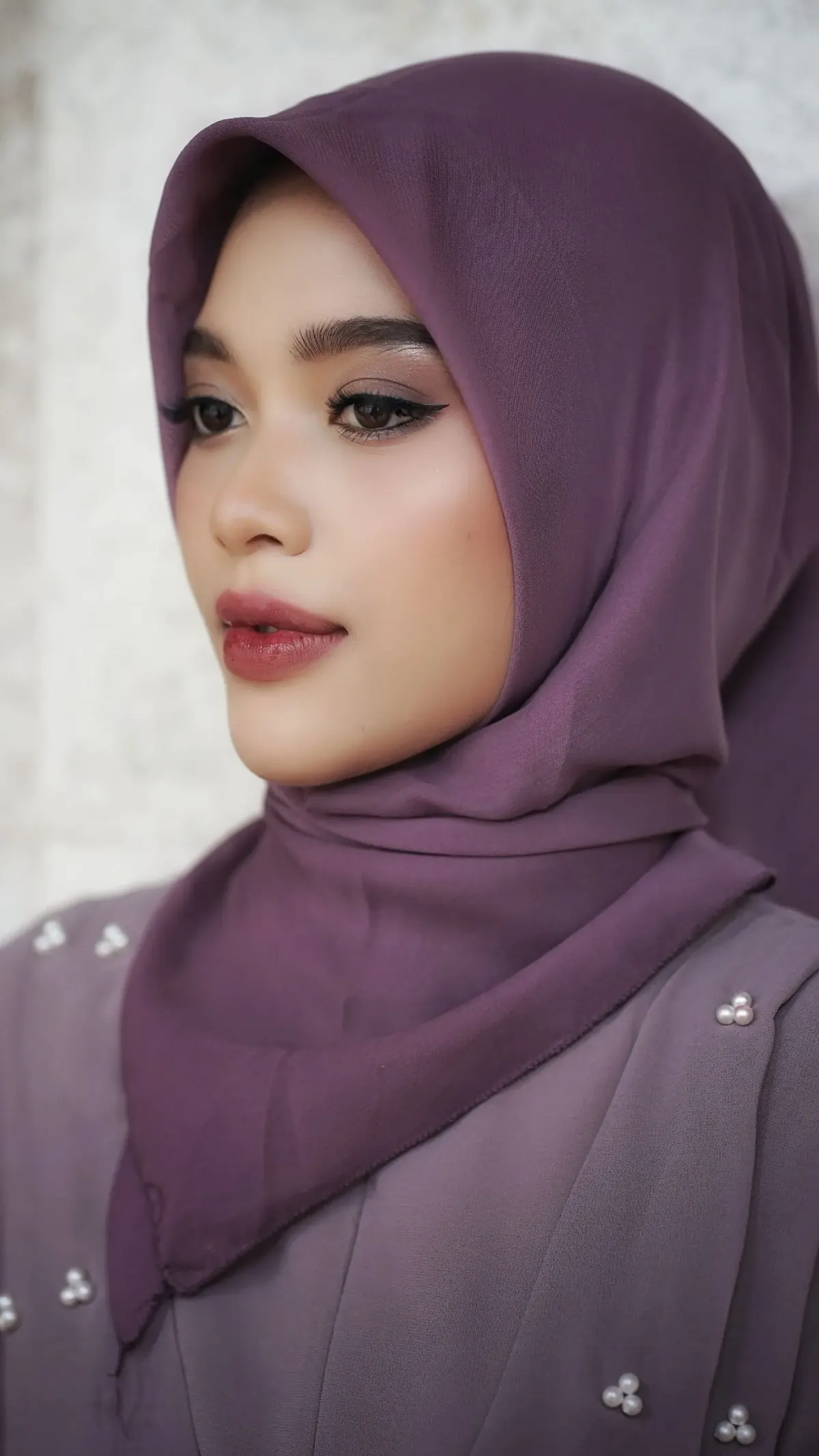 Potret samping perempuan berhijab segi empat ungu polos yang disematkan di bawah dagu dengan sudut kain jatuh di depan dada, memakai busana ungu muda berhias manik mutiara.