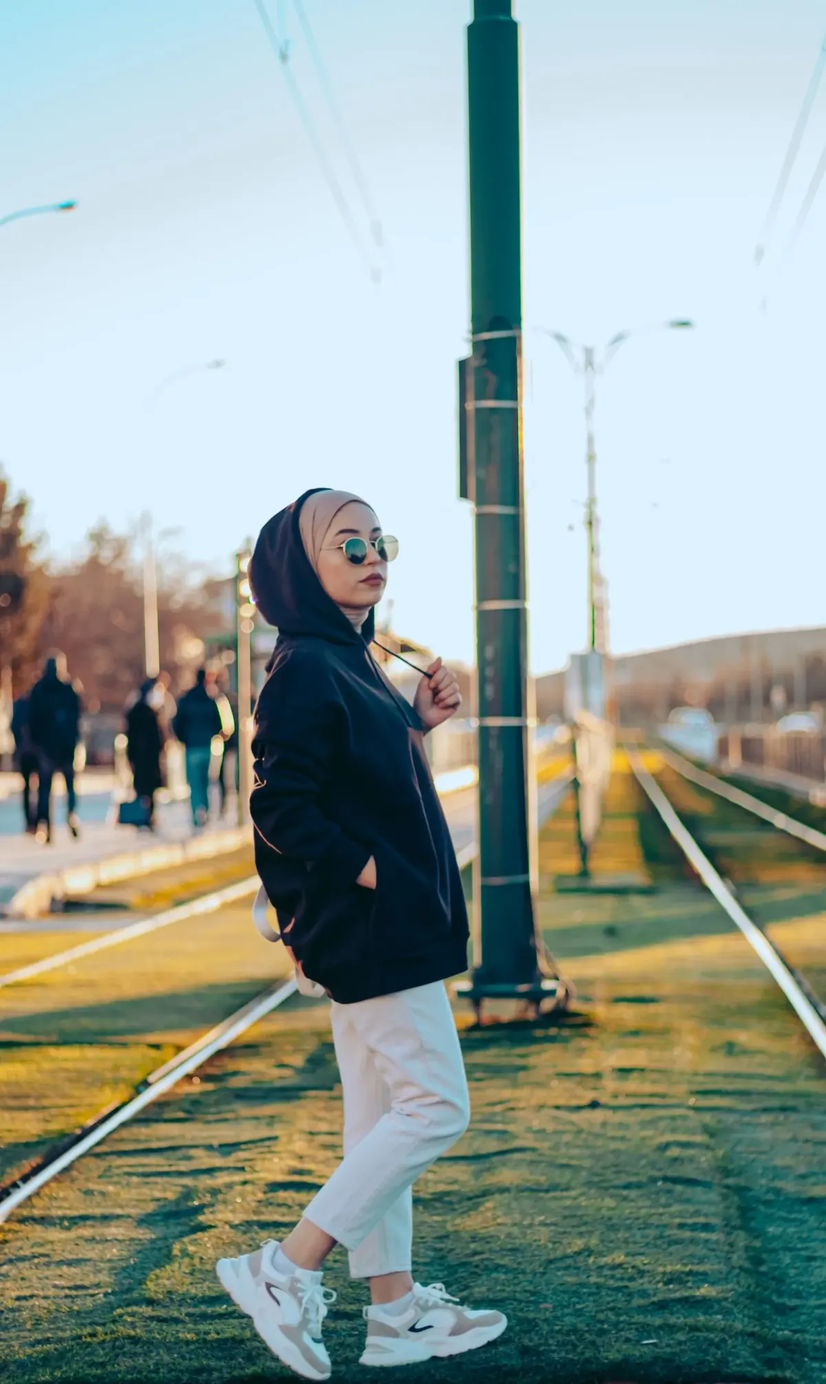 Perempuan berhijab krem dan berkacamata bulat memakai hoodie hitam, jins putih, dan sneakers putih abu-abu, berdiri di dekat rel trem kota.