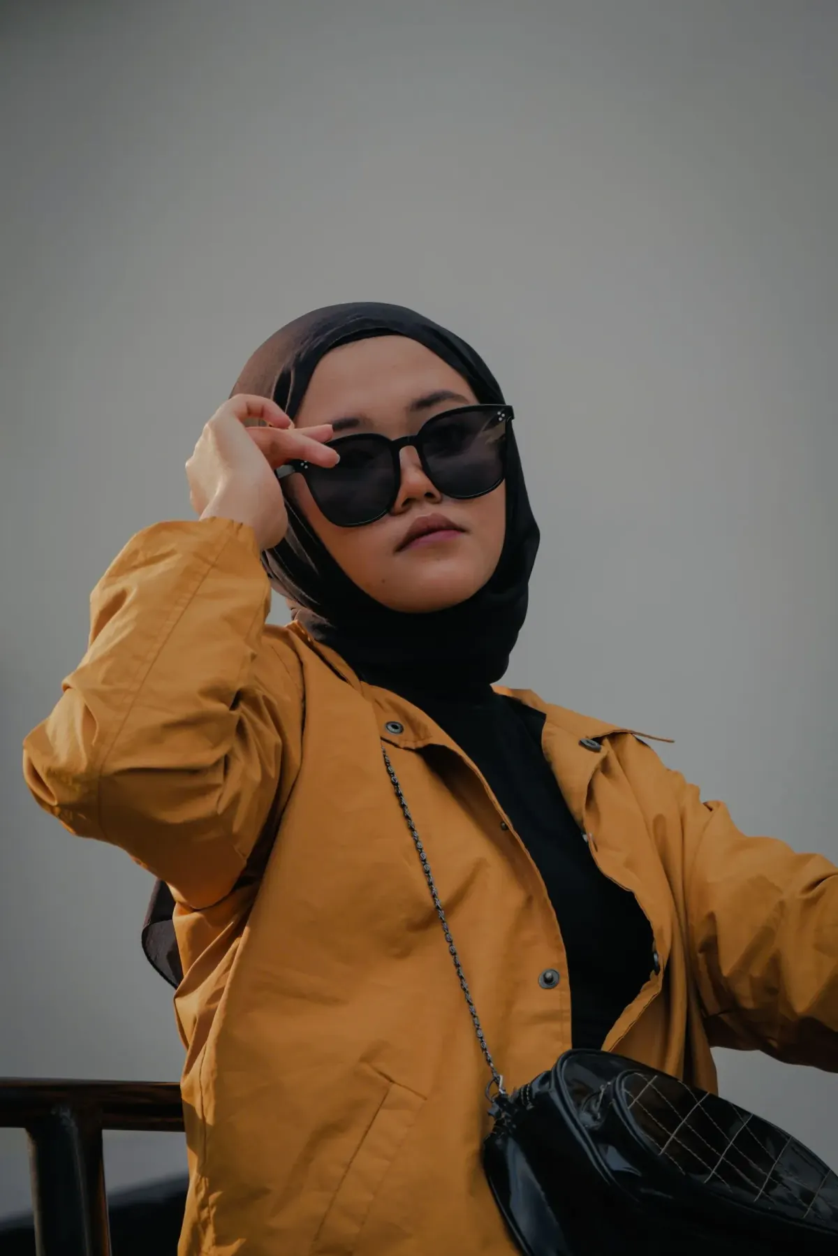 Perempuan berjilbab hitam dan atasan hitam memakai jaket kuning mustard berkancing, kacamata hitam, dan tas selempang hitam bertali rantai, berpose di depan dinding abu-abu.