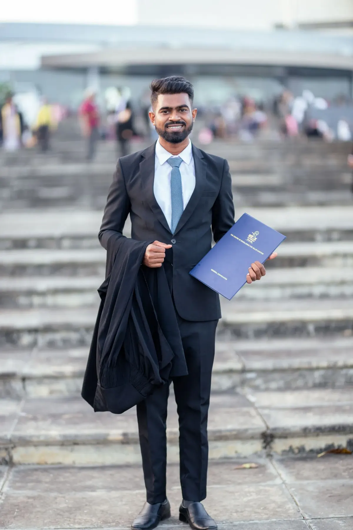 Pria memakai jas dan celana hitam, kemeja biru muda, dasi biru muda, dan sepatu hitam, memegang toga hitam dan map ijazah biru di tangga, Hambantota, Sri Lanka.