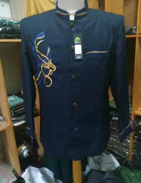 Jas koko navy berkerah berdiri dengan kancing logam, list kuning dan biru di kerah, saku, dan manset, serta bordir kaligrafi emas di dada kiri, dipajang di manekin dalam sebuah toko.