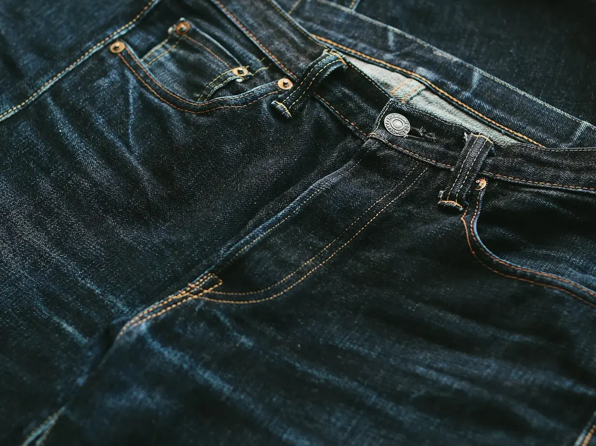Bagian depan celana jeans biru tua dengan garis-garis pudar terang di sekitar paha dan resleting akibat pemakaian.