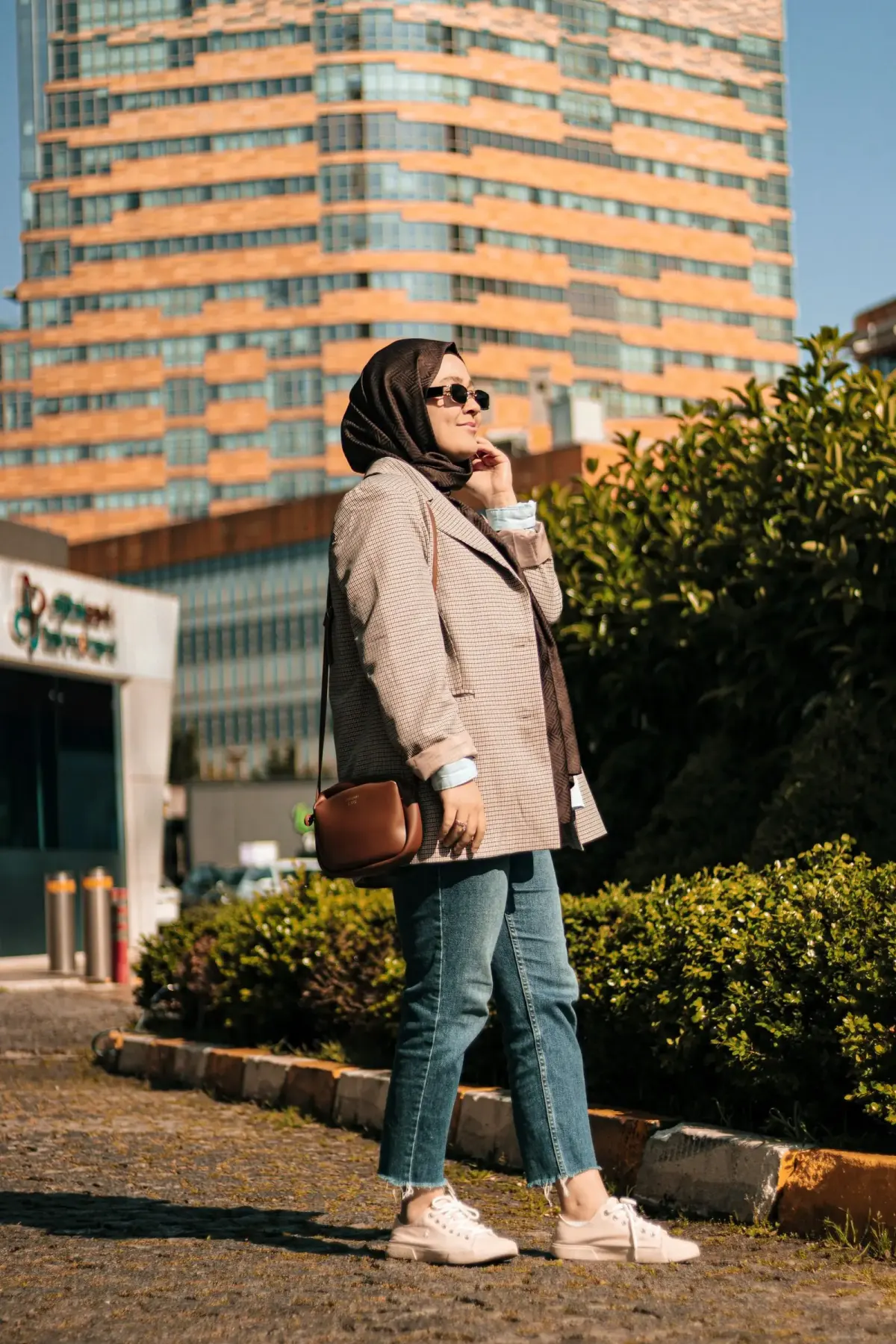 Perempuan berjilbab cokelat tua dan berkacamata hitam memakai blazer motif kotak kecil warna beige cokelat, celana jeans biru potongan lurus sebatas mata kaki dengan ujung berserabut, tas selempang cokelat, dan sepatu kets krem.