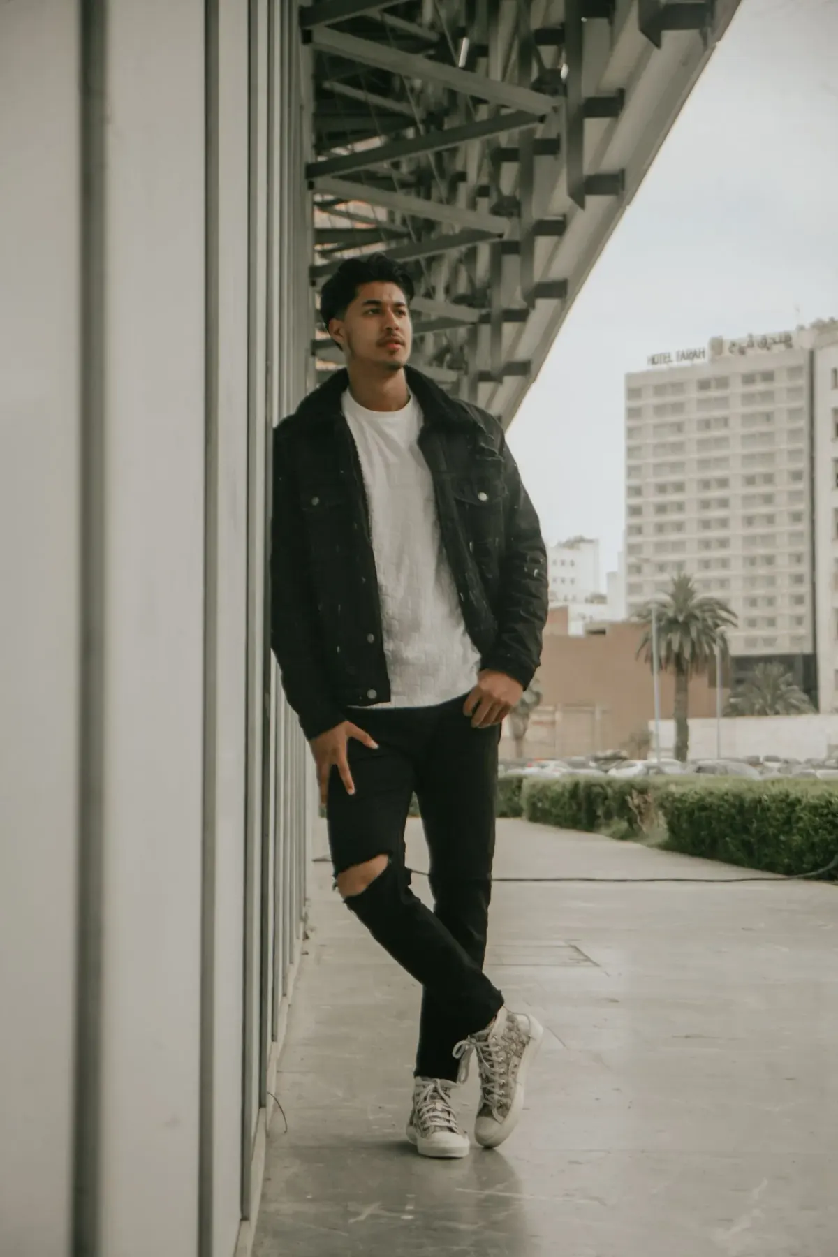 Pria bersandar di dinding gedung memakai jaket denim hitam, kaus putih bertekstur, celana jeans hitam ramping dengan sobekan di lutut, dan sepatu kets bertali warna abu-abu.