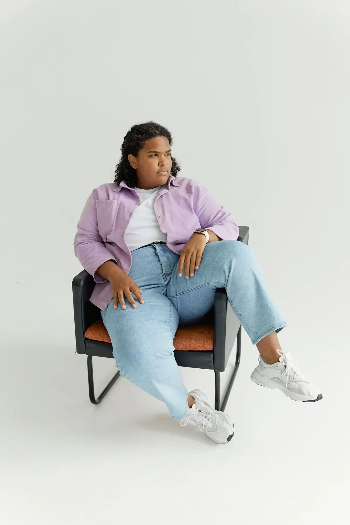 Perempuan duduk di kursi studio memakai kemeja lilac yang dibiarkan terbuka di atas kaus putih, celana jeans biru muda berpotongan lurus, dan sepatu kets putih tebal.