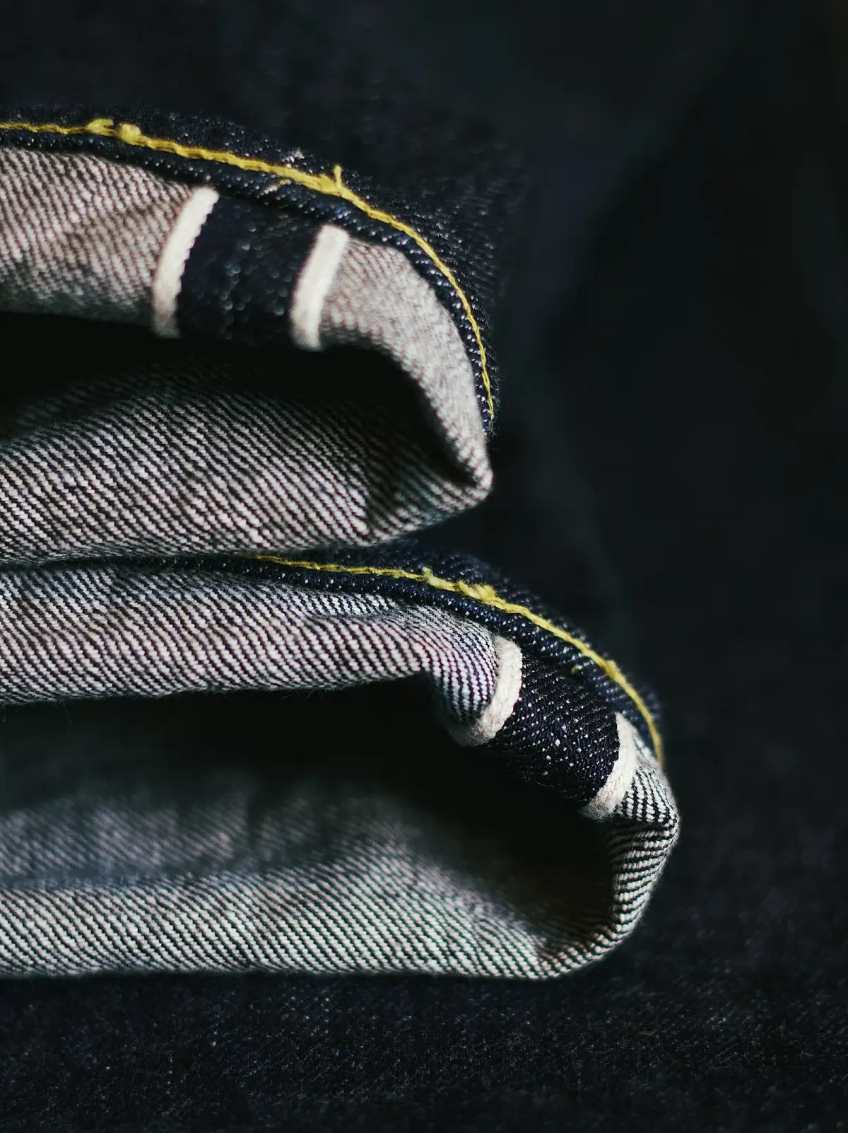 Ujung kaki celana jeans biru tua yang digulung dua kali sehingga tepi kain selvedge berupa garis putih dengan jahitan kuning terlihat di bagian dalam.