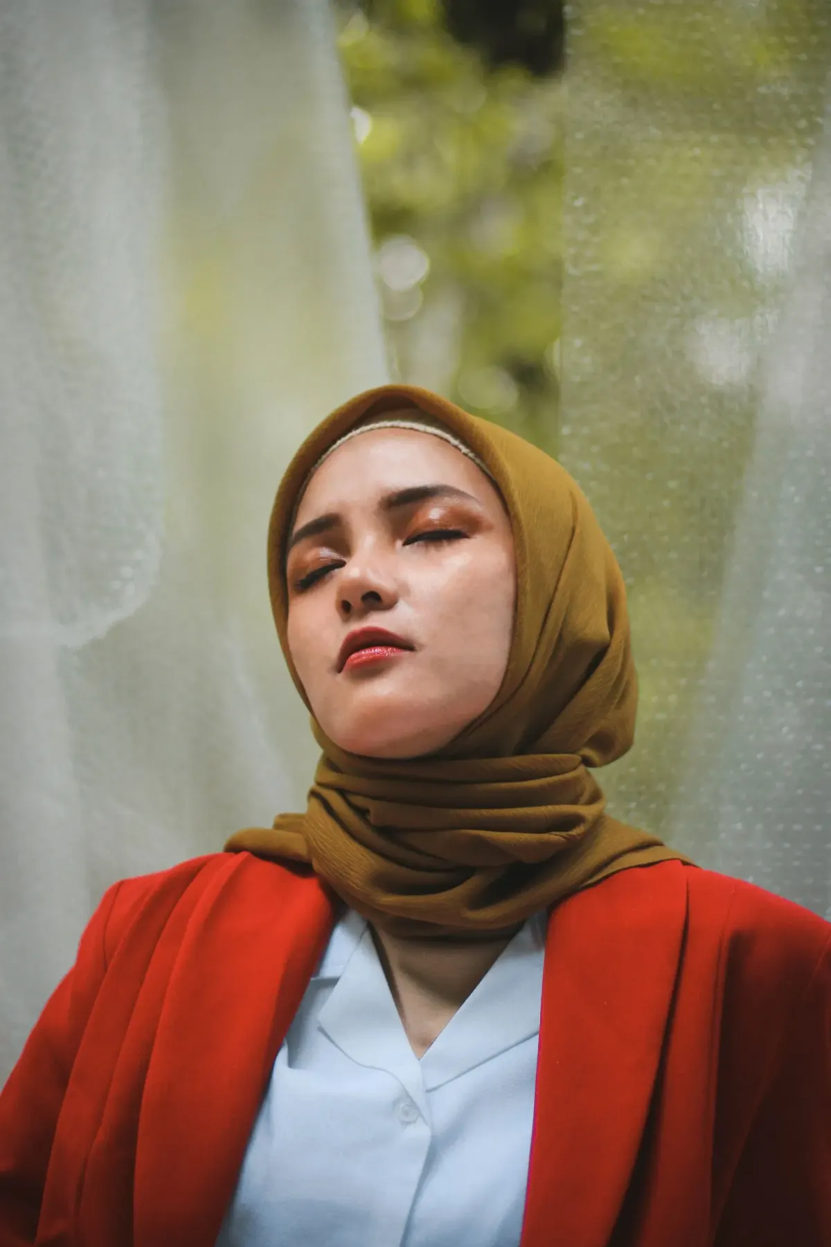 Perempuan berjilbab cokelat keemasan yang dililit ke leher, memakai kemeja biru muda dan blazer merah, berdiri di antara tirai putih tipis.
