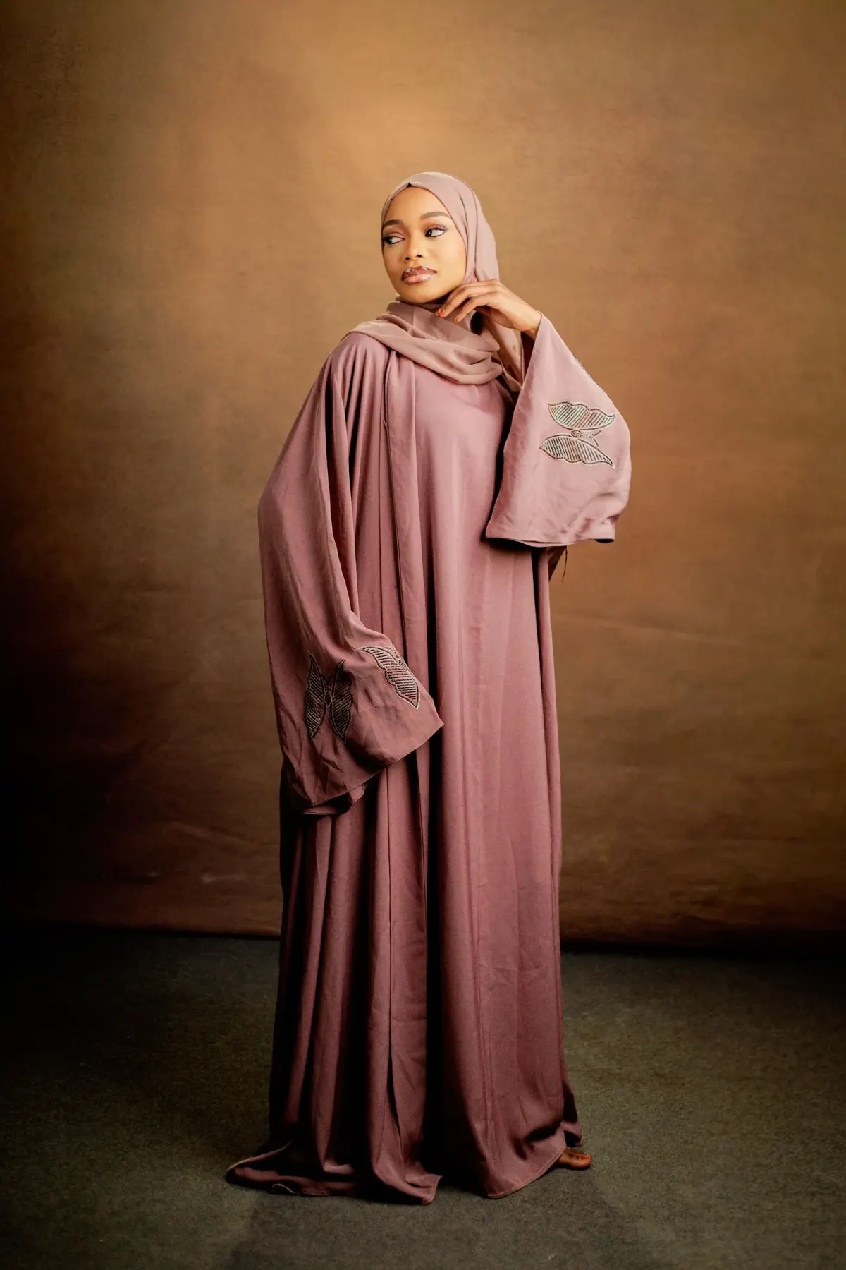 Perempuan berhijab mauve memakai busana panjang mauve berpotongan sangat longgar dengan lengan lebar berhias aplikasi bermotif kupu-kupu, berdiri di depan latar studio cokelat.