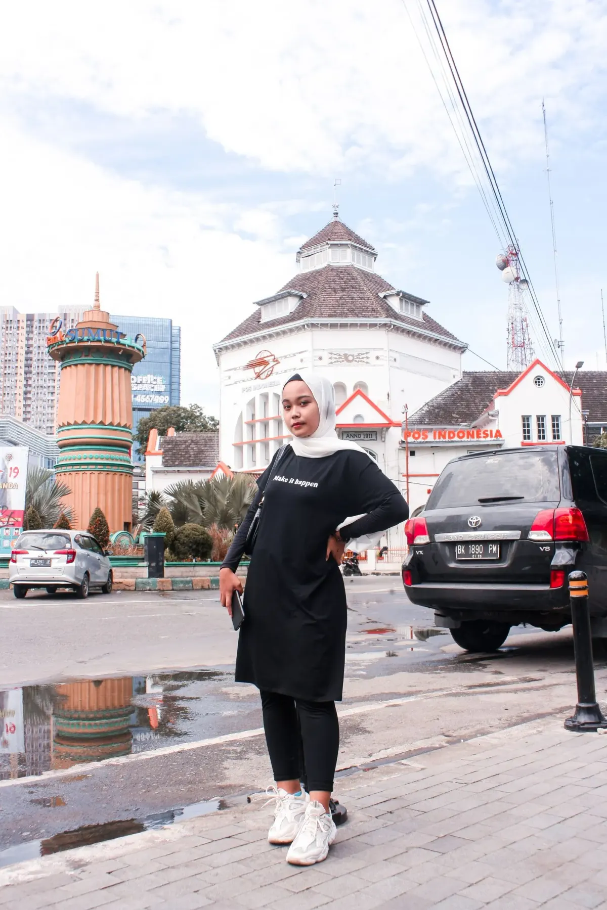Perempuan berhijab putih memakai kaus panjang hitam bertulisan, legging hitam, dan sneakers putih, berpose di trotoar depan gedung Kantor Pos Indonesia bergaya kolonial.
